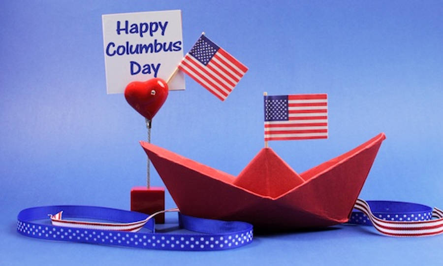 Columbus Day 桌布 壁紙 — 一艘紅色紙船，上面有美國國旗，在藍色絲帶波浪上行駛，旁邊有一個心形氣球和一個「哥倫布日快樂」標誌。