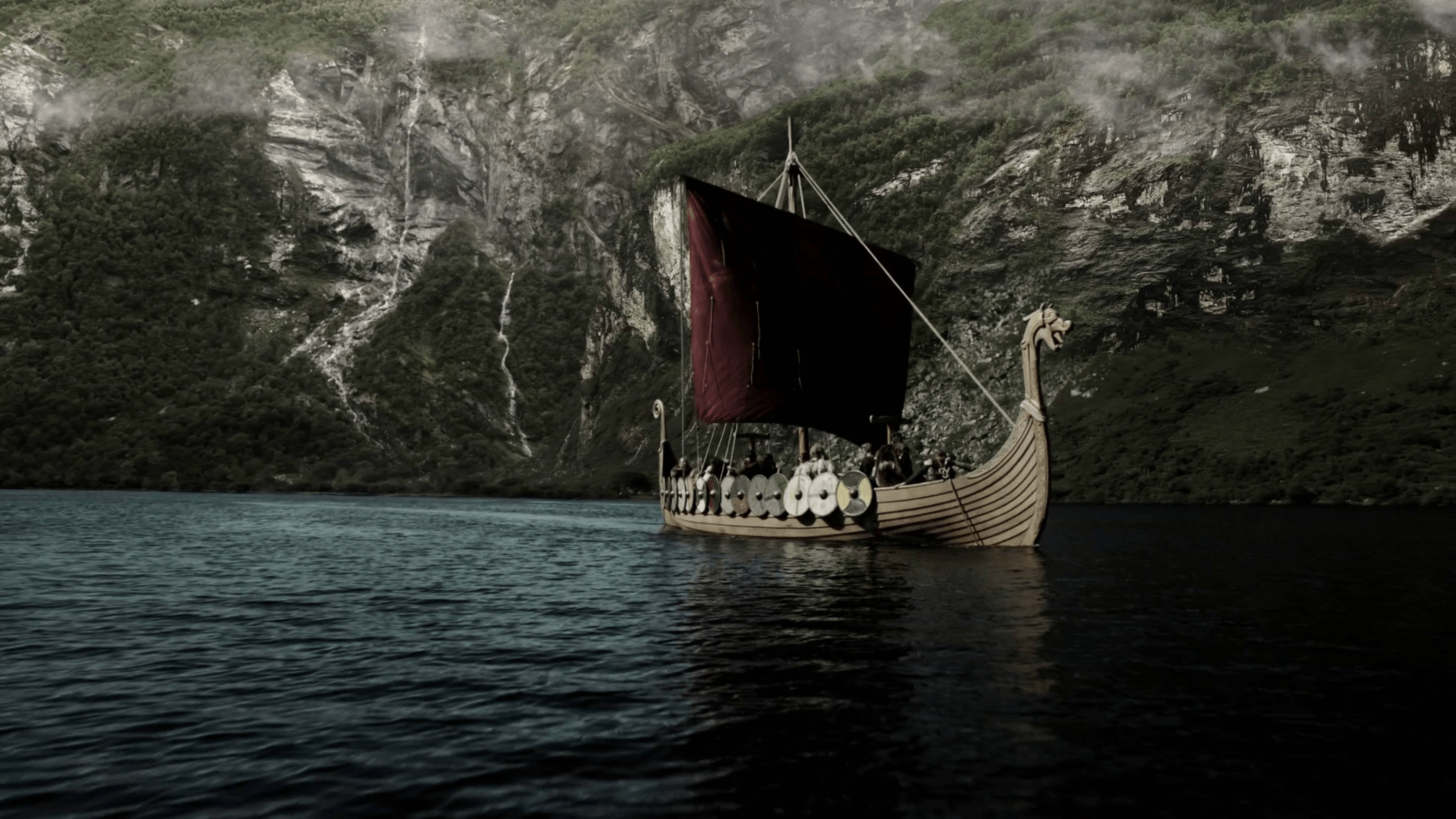 Viking Ship 桌布 壁紙 — 一艘維京長船，掛著深色帆，在平靜的水面上滑行，兩旁是高聳的霧氣繚繞的山脈。