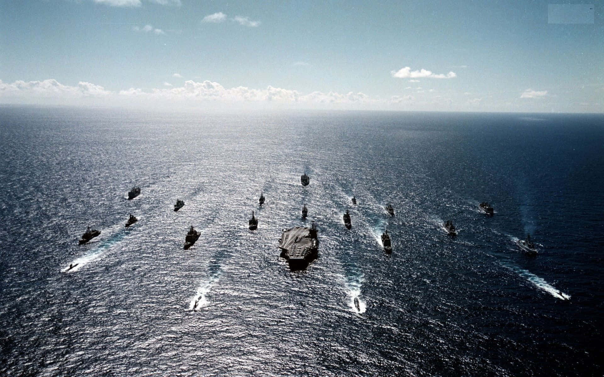 美國 桌布 壁紙 — 一艘航空母艦帶領一支軍艦艦隊在部分多雲的天空下，編隊航行在廣闊的藍色海洋上。