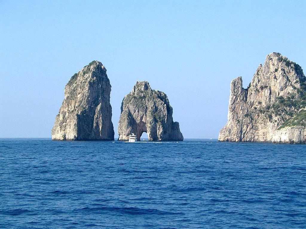Capri 桌布 壁紙 — 一艘船在晴朗的天空下航行穿過法拉廖尼岩石從深藍色海中升起的標誌性天然拱門。