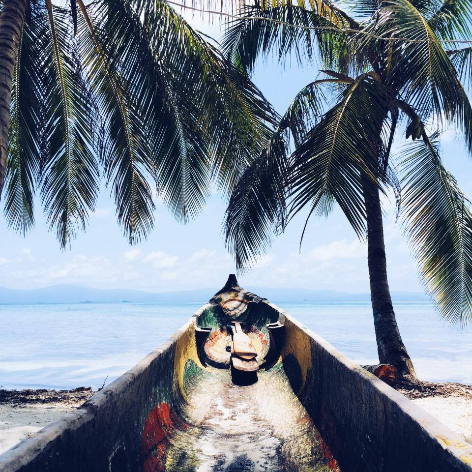 Tropical Beach 桌布 壁紙 — 一艘色彩繽紛的彩繪獨木舟停泊在沙灘上，搖曳的棕櫚樹下，平靜的海洋和遠山在明亮的天空下。