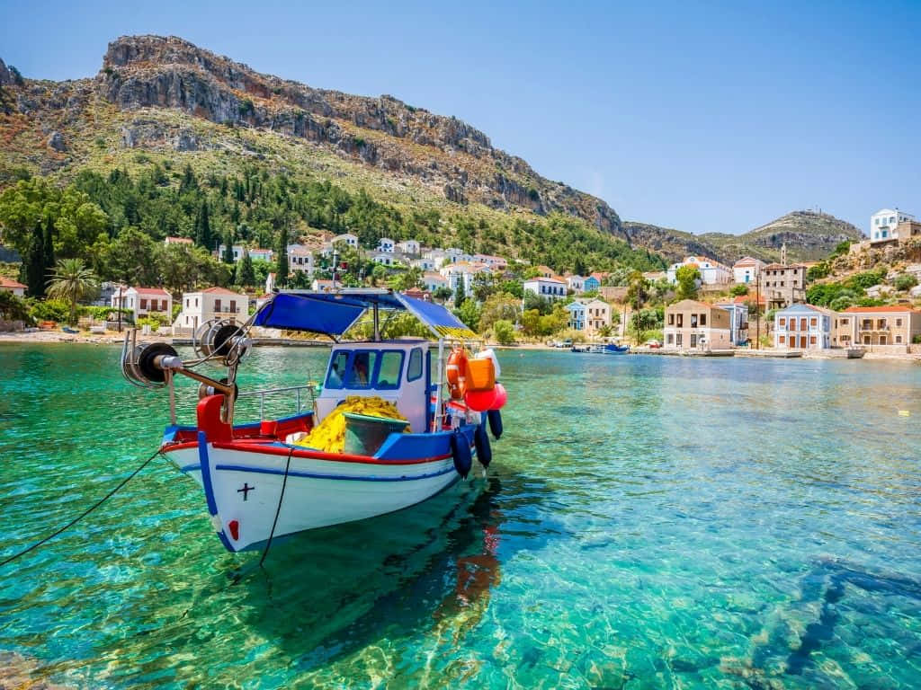Greek Island 桌布 壁紙 — 一艘色彩繽紛的漁船漂浮在清澈碧綠的海水中，旁邊是山坡村莊，白色建築物在明亮的藍天下。