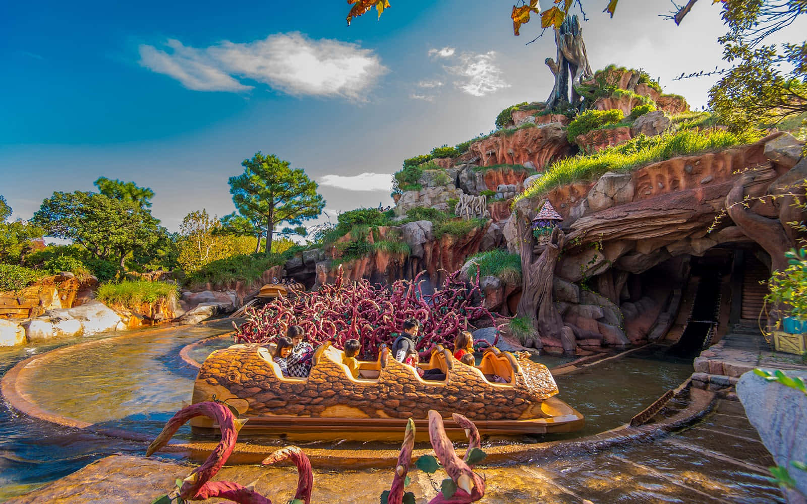 Splash Mountain 桌布 壁紙 — 一艘色彩鮮豔的水上遊樂船載著乘客，在岩石嶙峋、叢林主題的航道上航行，天空湛藍，白雲點綴。