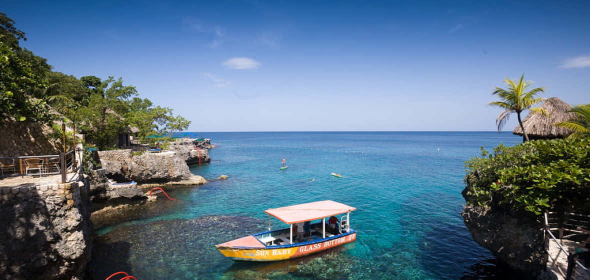 Jamaican Island 桌布 壁紙 — 一艘色彩鮮豔的玻璃底船在明亮的藍天下，穿梭於鬱鬱蔥蔥的綠色懸崖之間，滑行在清澈見底的碧綠海水上。