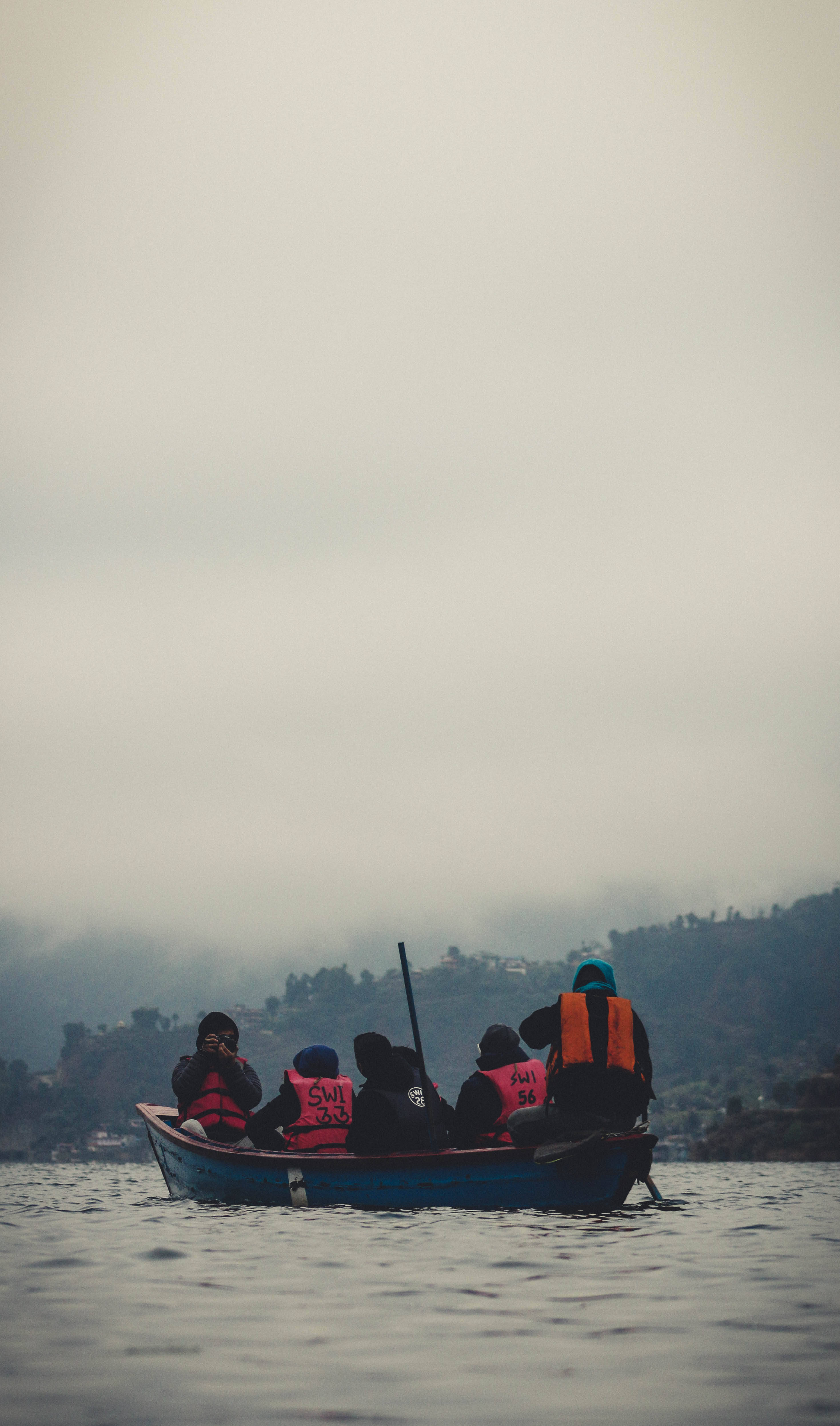 Nepal 桌布 壁紙 — 一艘藍色小船載著幾位穿著紅色救生衣的人，穿越霧氣瀰漫的湖泊，背景是朦朧的山丘。