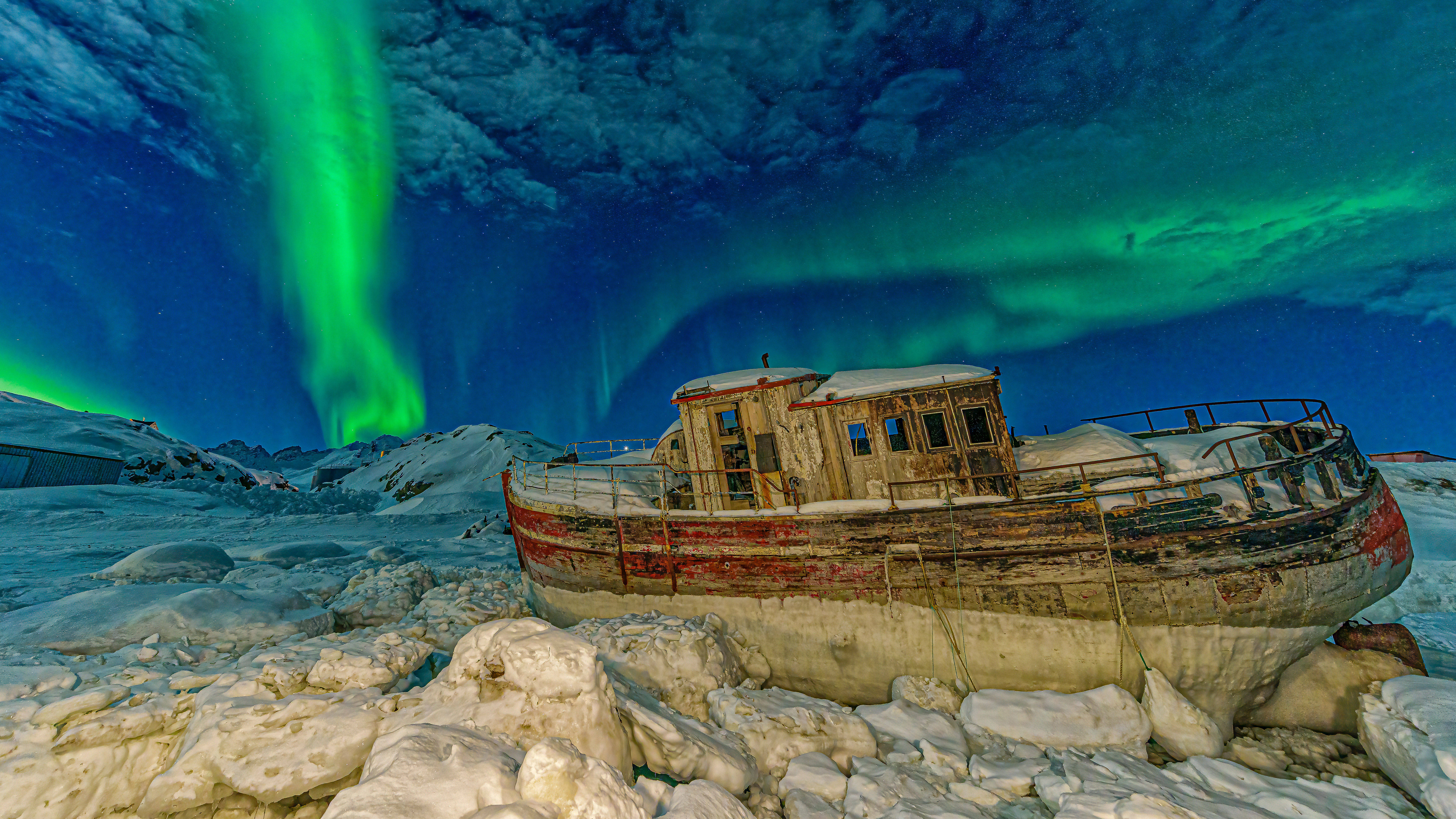 Greenland 桌布 壁紙 — 一艘被遺棄的木船停泊在雪覆蓋的冰面上，上方是 vibrant 綠色極光在繁星點點的夜空中舞動。