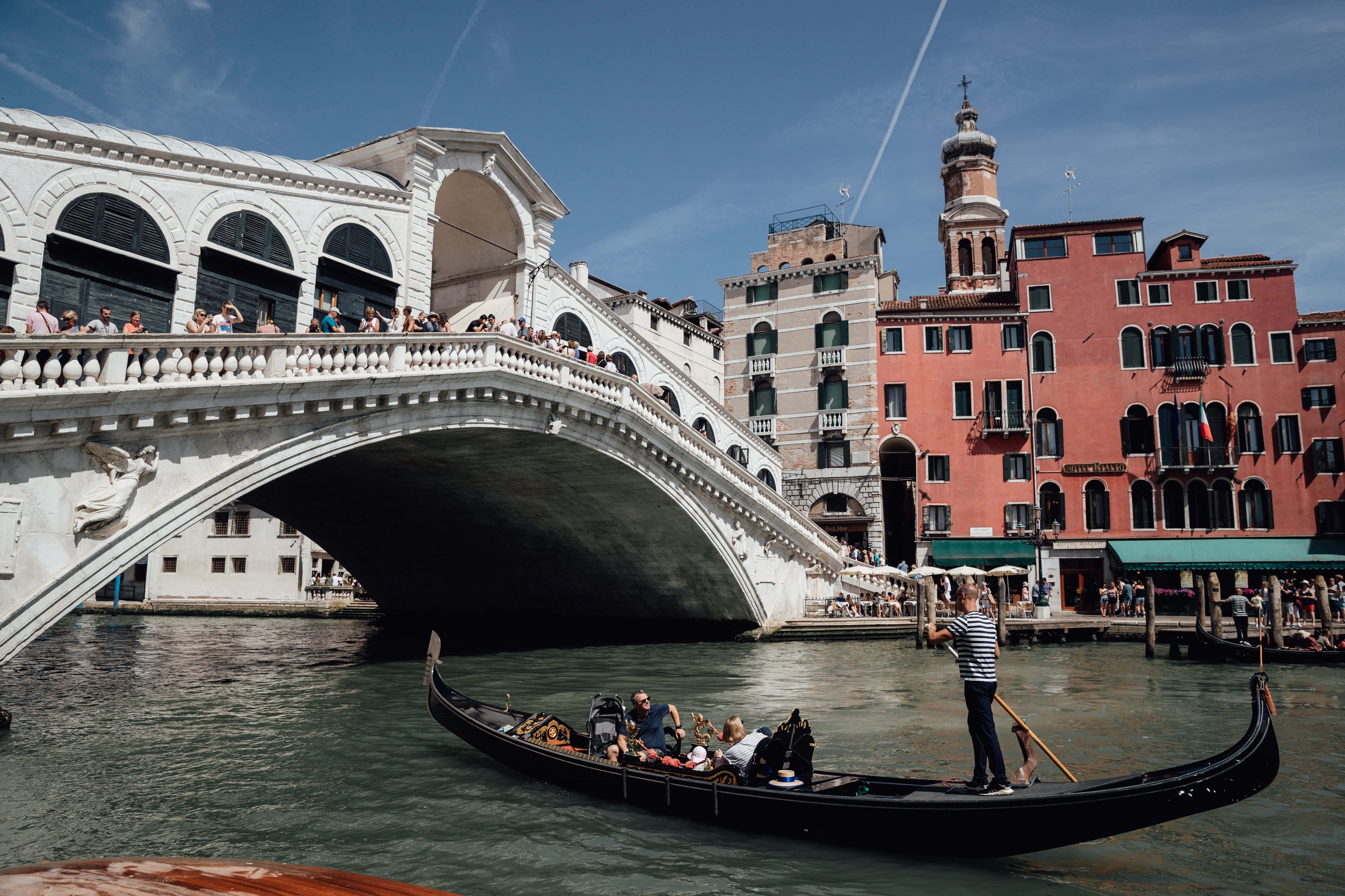 venice 桌布 壁紙 — 一艘貢多拉在威尼斯標誌性的里亞爾托橋下滑行，遊客在晴朗的藍天下欣賞運河景色。