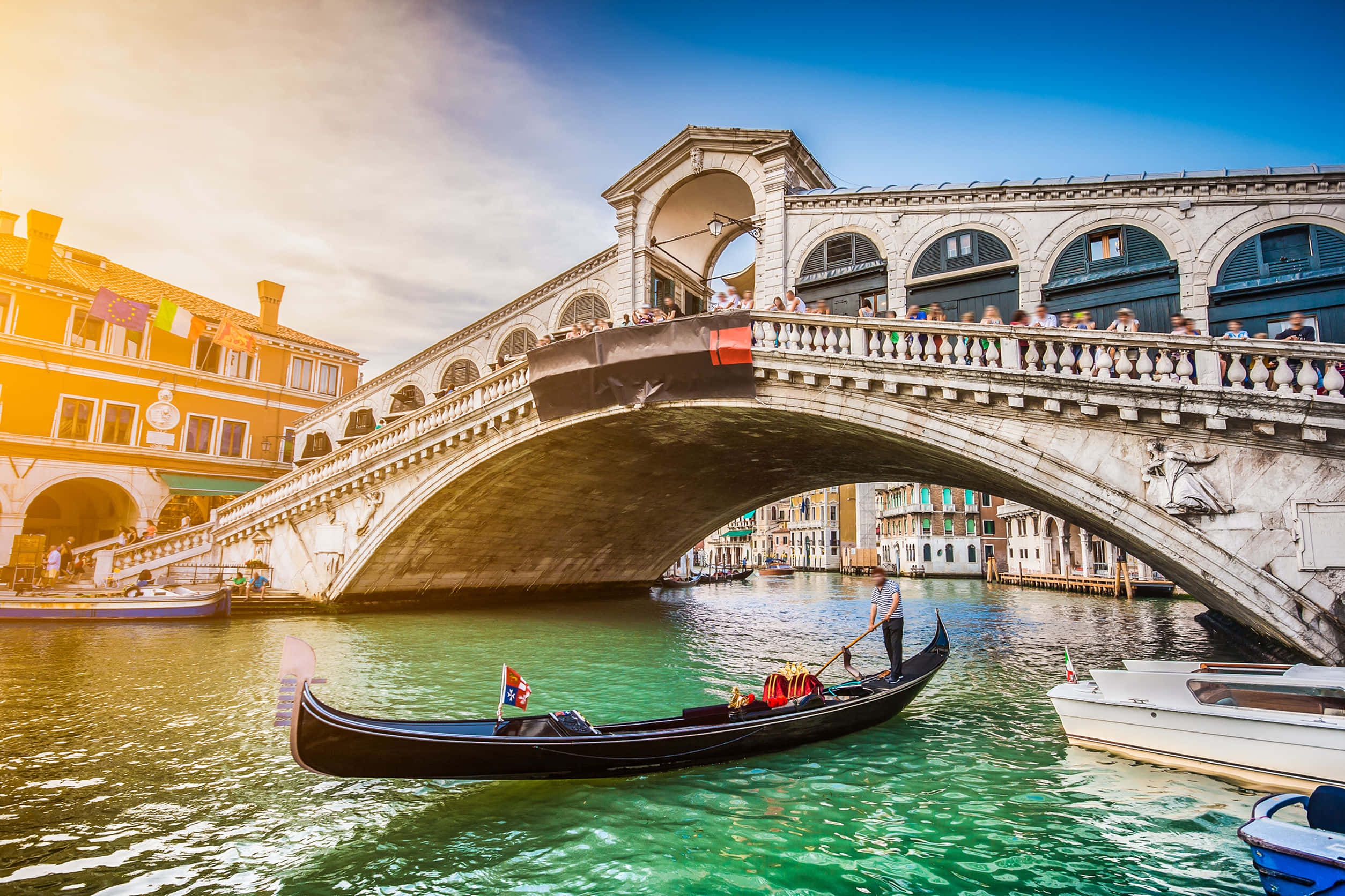 venice 桌布 壁紙 — 一艘貢多拉在威尼斯標誌性的里亞爾托橋下滑行，遊客擠滿橋上，色彩繽紛的建築物沿運河排列。