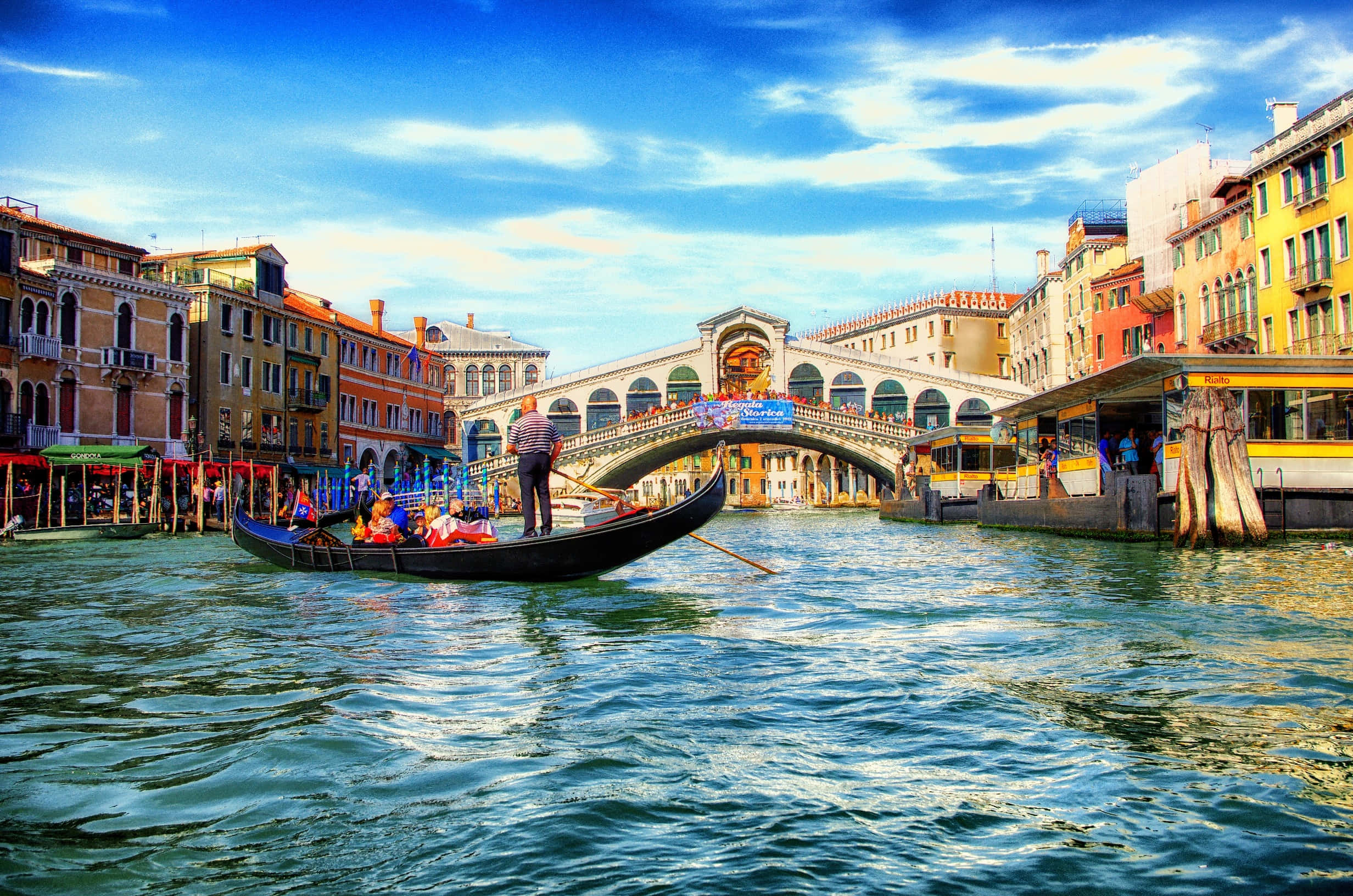 venice 桌布 壁紙 — 一艘貢多拉在威尼斯標誌性的里亞爾托橋下滑行，色彩繽紛的建築物沿著運河排列，頭頂是明亮的藍天。