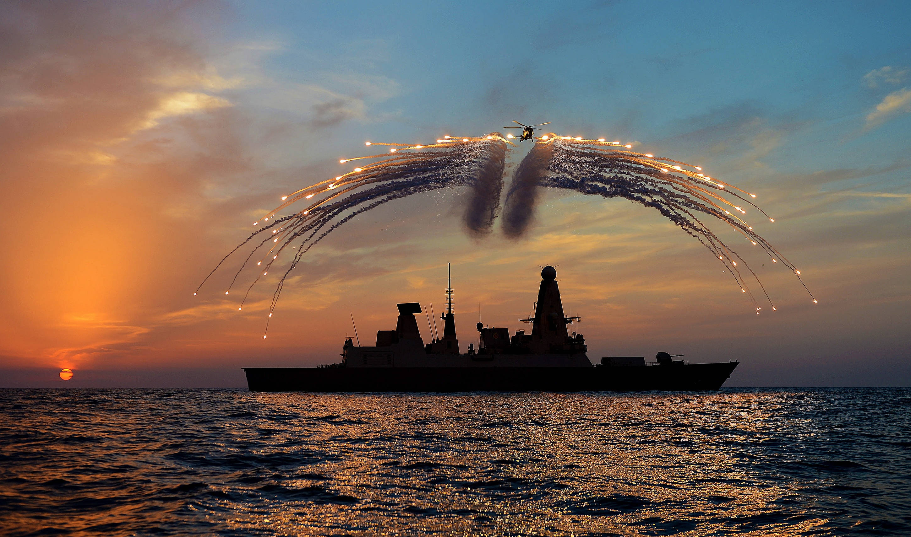 Helicopter 桌布 壁紙 — 一艘軍艦在火紅的夕陽下呈現剪影，直升機在海洋上空以戲劇性的弧線釋放照明彈。
