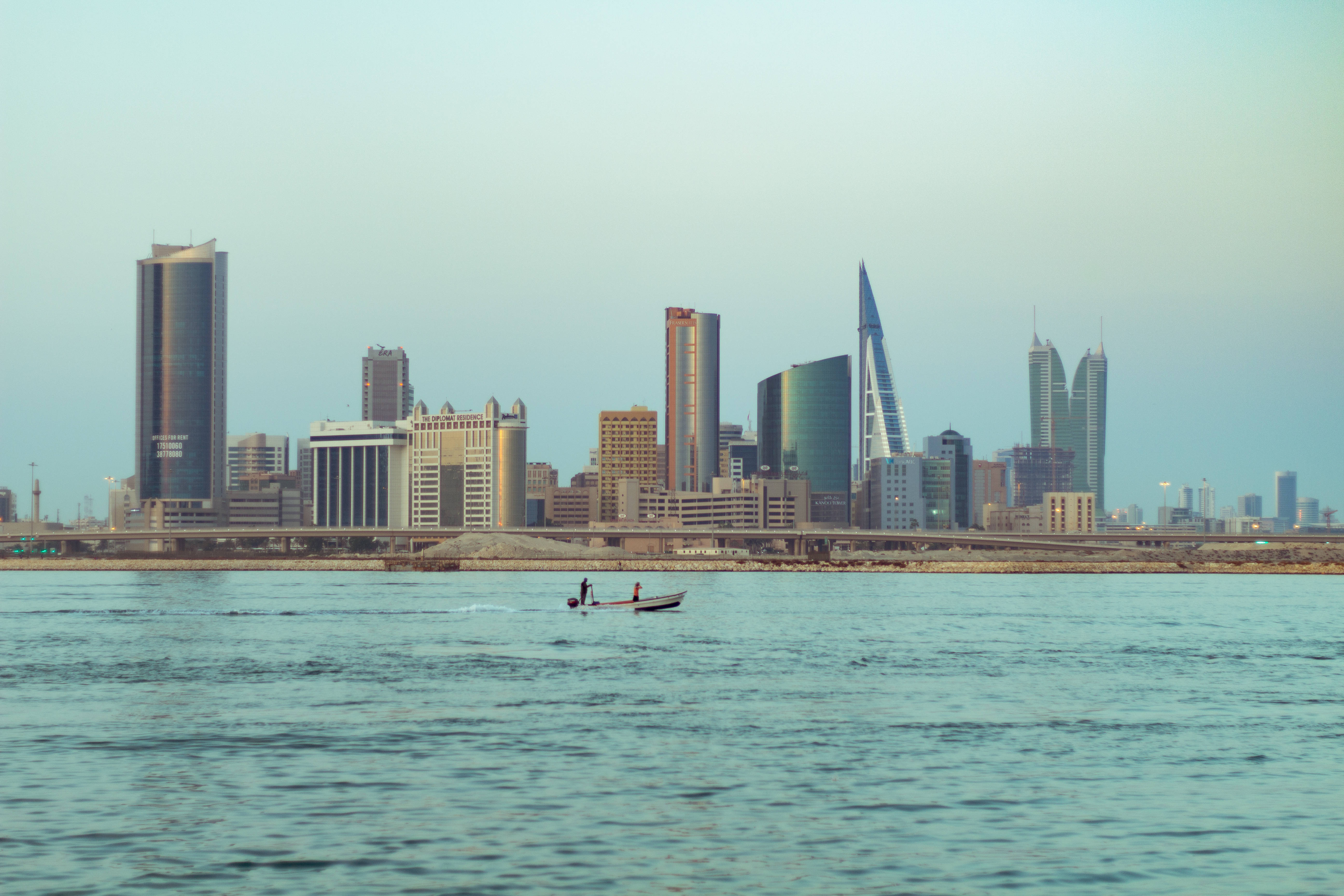 Bahrain 桌布 壁紙 — 一艘載有兩人的小船在黃昏時分，於平靜的藍色水面上滑行，前方是現代化的城市天際線。