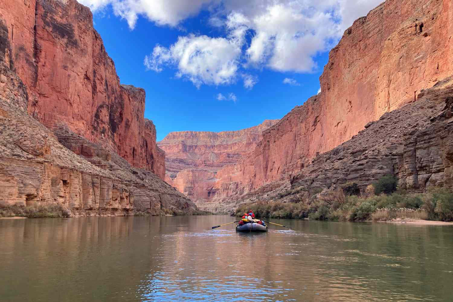 grand canyon 桌布 壁紙 — 一艘載有冒險家的木筏在狹窄的峽谷中滑行，兩旁是高聳的紅色岩壁，上方是明亮的藍天和蓬鬆的白雲。