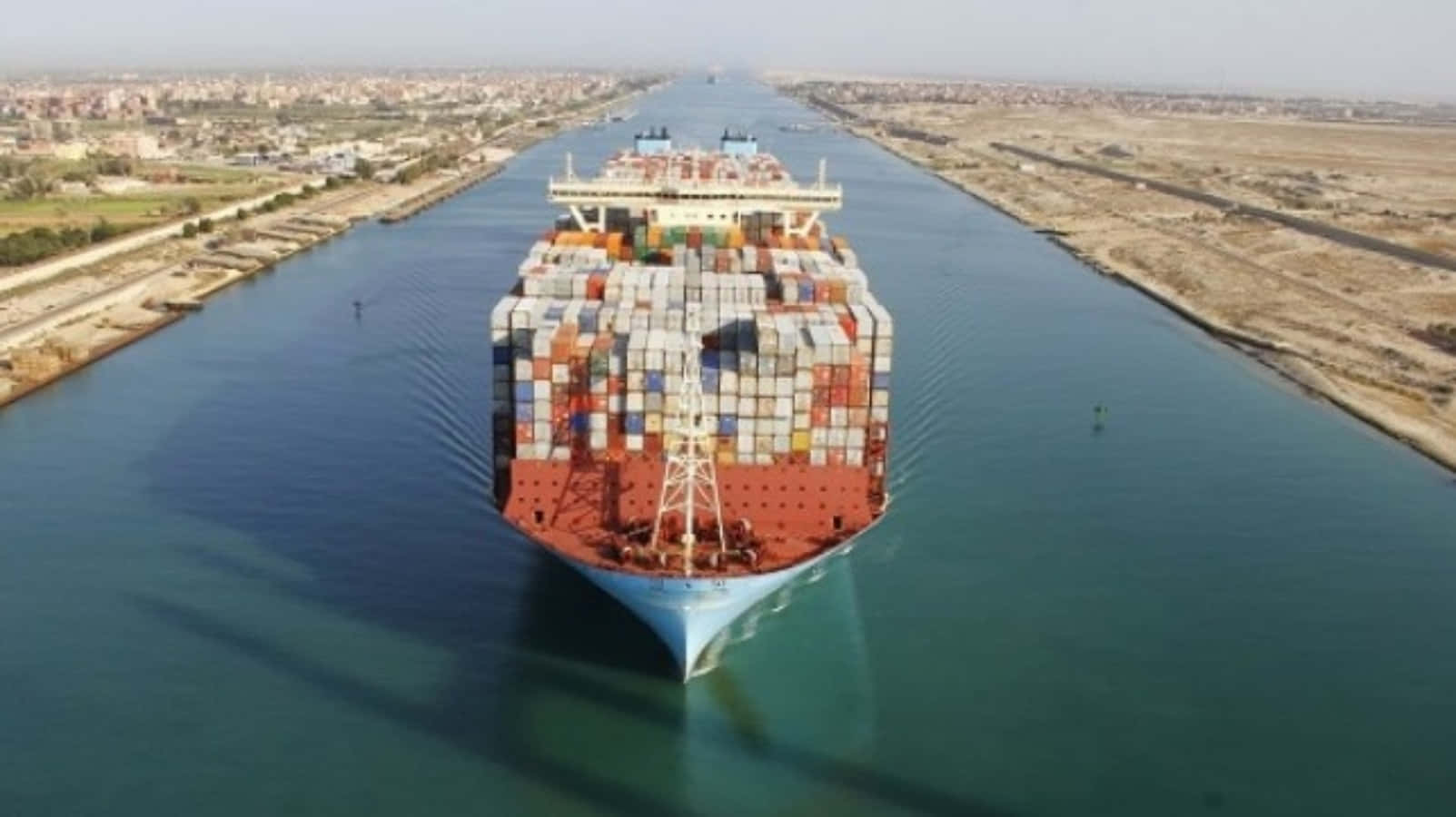 Cargo Ship 桌布 壁紙 — 一艘載滿彩色貨物的巨型貨櫃船在寬闊的人工運河中航行，運河兩旁是乾旱的土地和遠處的城市擴展區。