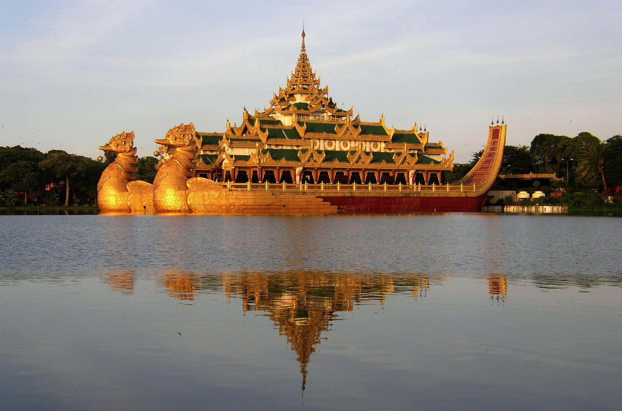 Yangon 桌布 壁紙 — 一艘金色華麗寺廟船在平靜水面上悠然漂浮，在柔和夕陽下呈現完美倒影。