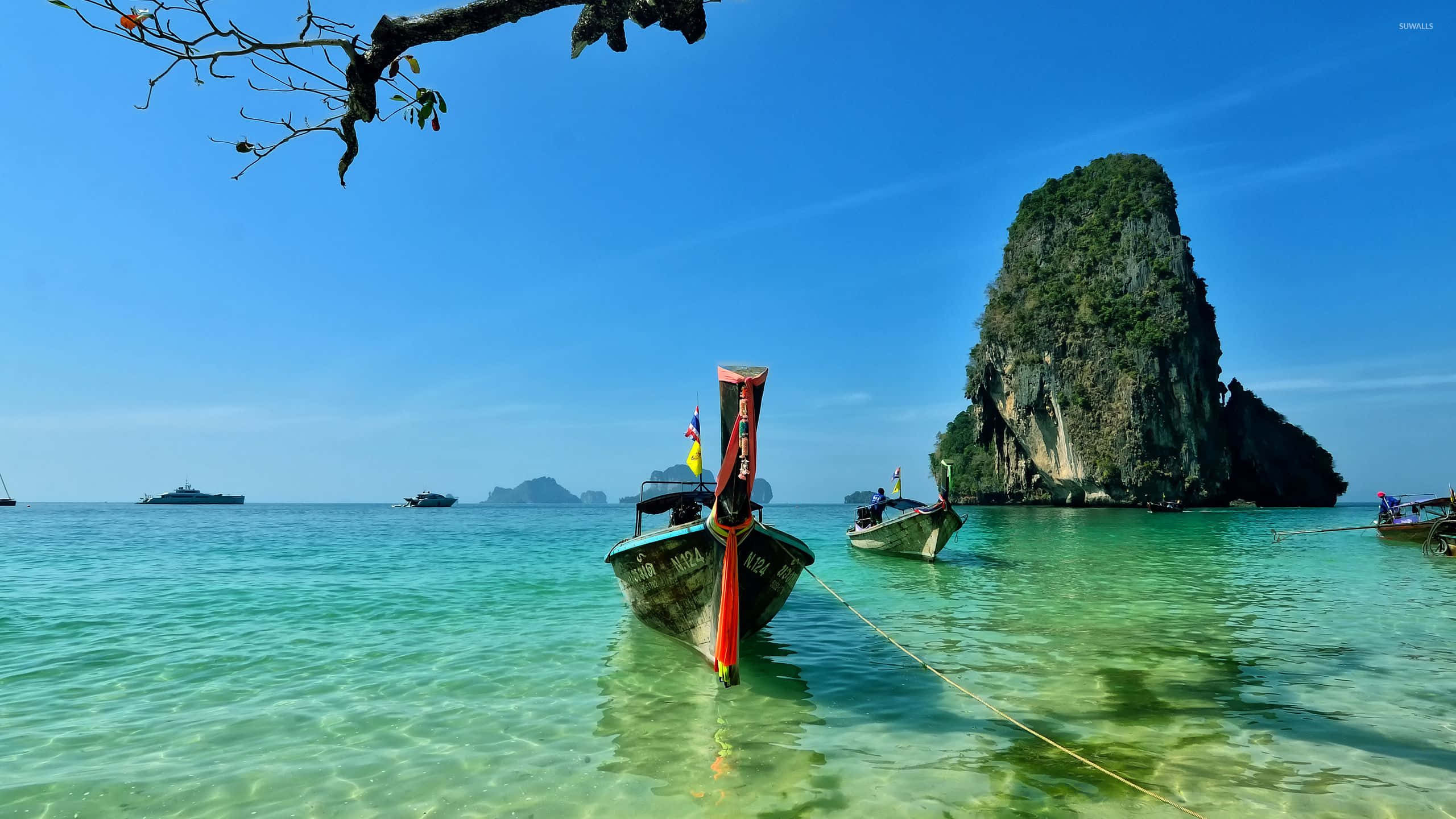 Thailand Beach 桌布 壁紙 — 一艘長尾船在明亮的藍天下，停泊在靠近一座高聳的叢林覆蓋的石灰岩島嶼的清澈碧綠海水中。