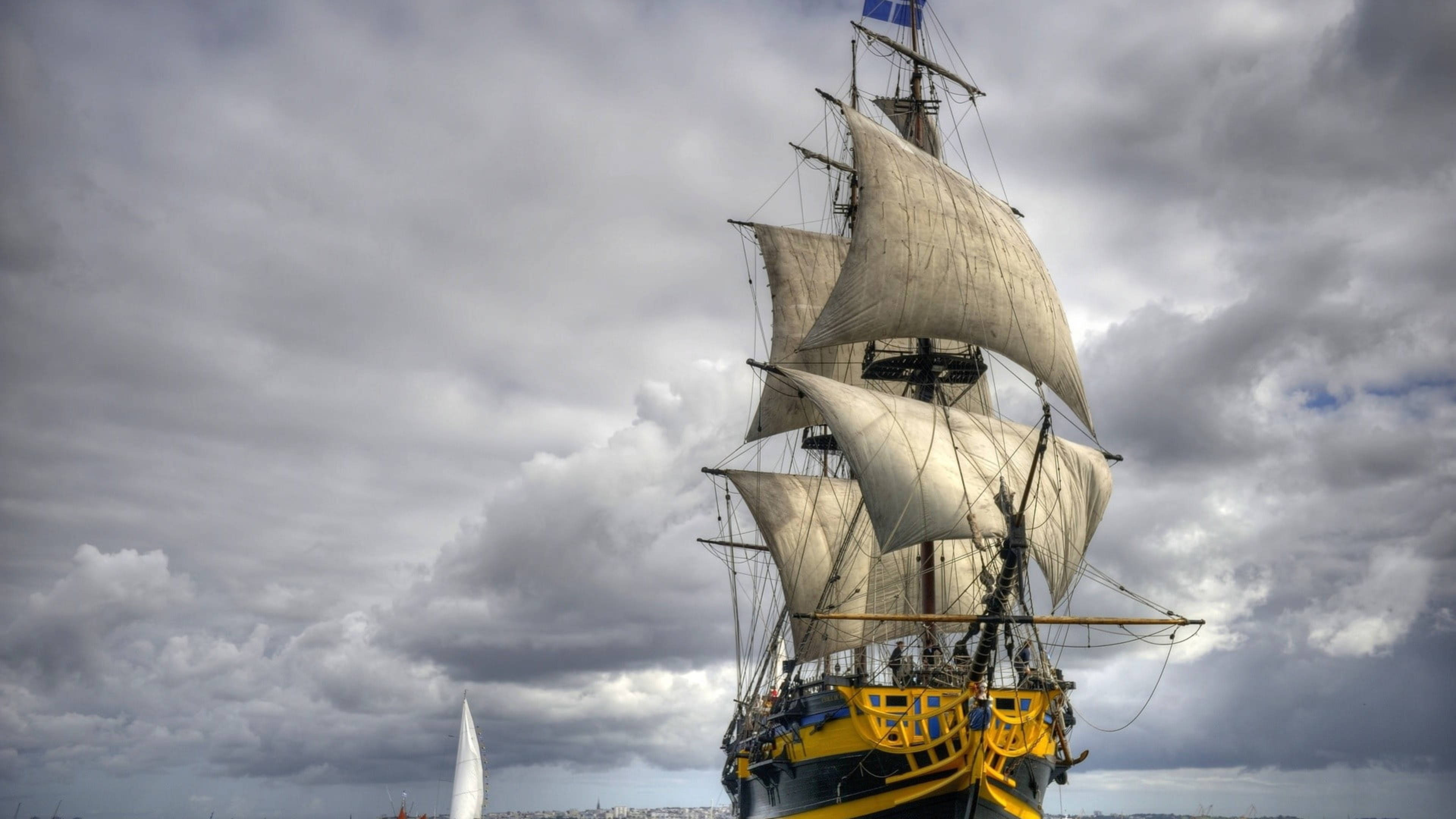 Sailing Ship 桌布 壁紙 — 一艘雄偉的大帆船，白色船帆鼓滿風，劃過海岸城市上空戲劇性的多雲天空。