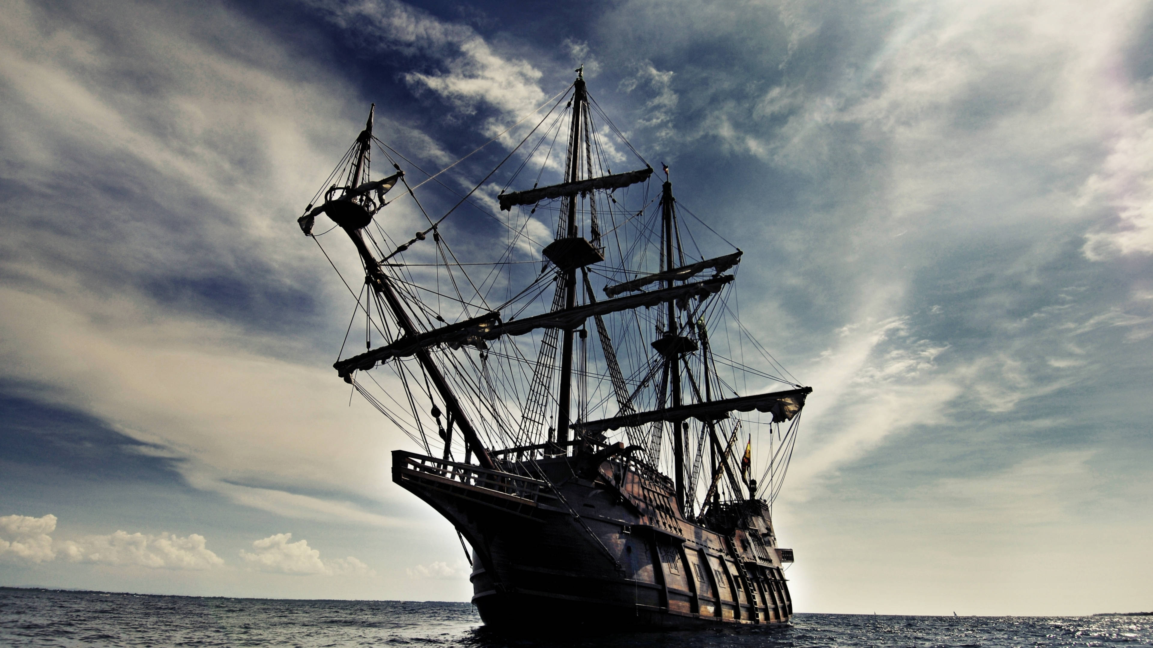 Pirates Of The Caribbean 桌布 壁紙 — 一艘雄偉的海盜船在戲劇性的多雲天空下航行，陽光穿透雲層。