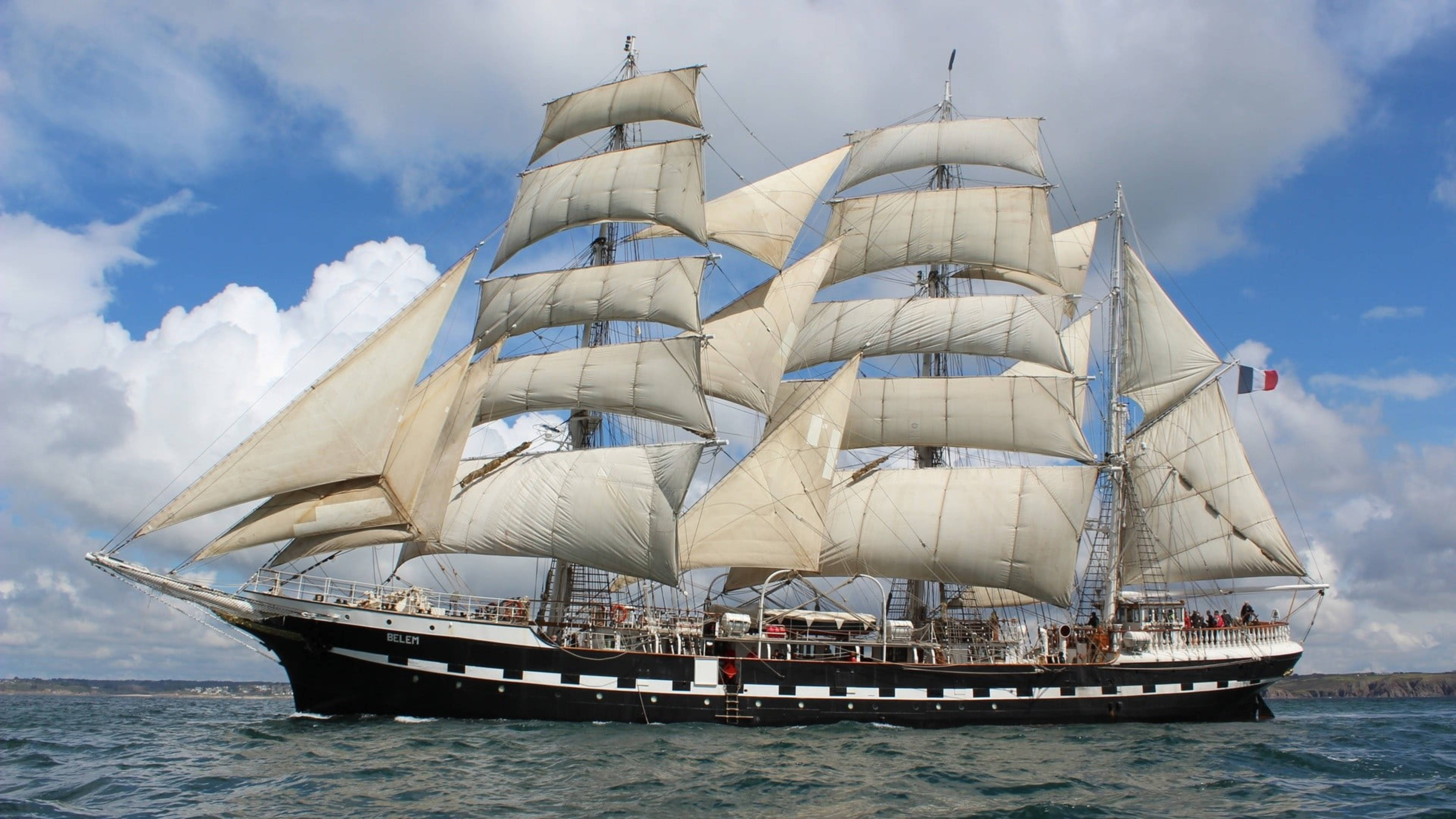 Sailing Ship 桌布 壁紙 — 一艘雄偉的高桅帆船，白色船帆隨風飄揚，在部分多雲的天空下滑過藍色水域。