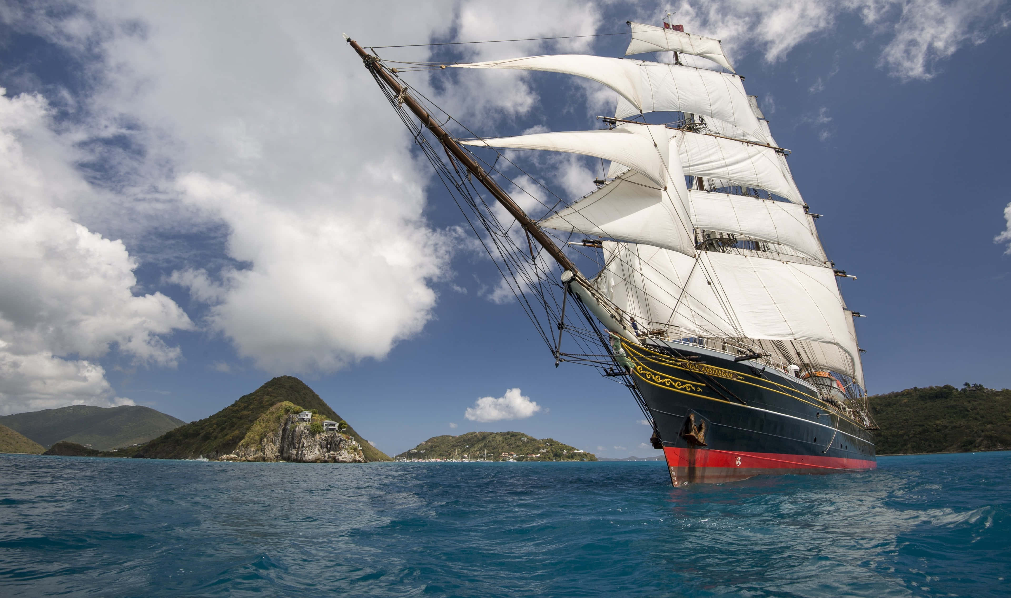 Sailboat 桌布 壁紙 — 一艘雄偉的高桅帆船，鼓著白色風帆，在部分多雲的藍天下，滑行於鬱鬱蔥蔥的綠色島嶼附近的碧綠海水之中。