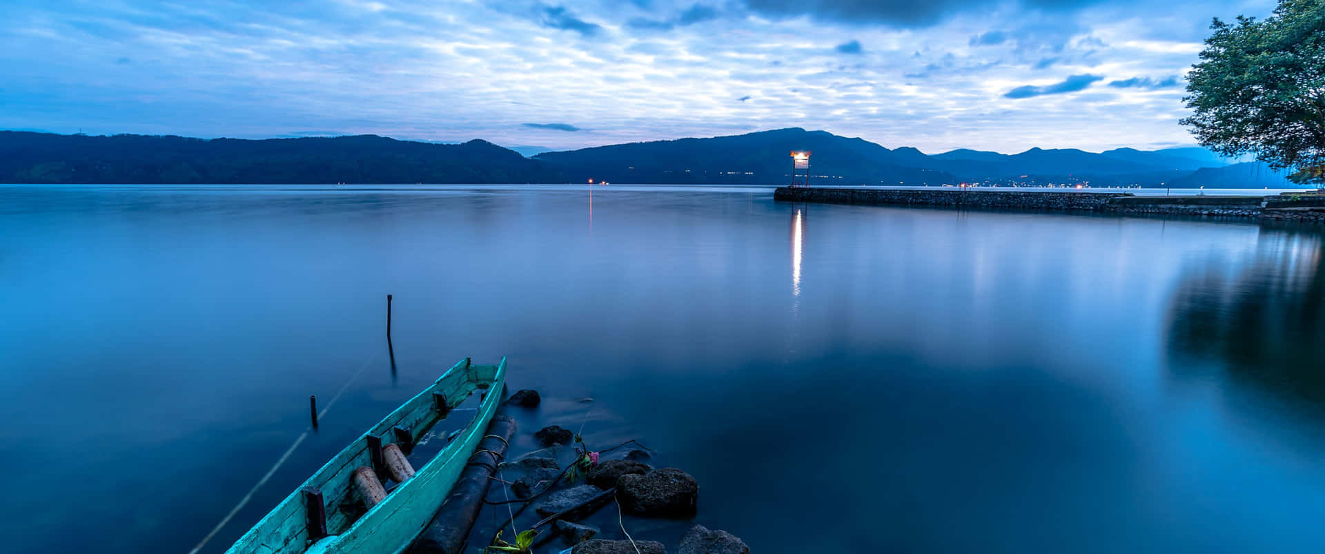 黃昏時岩石海岸上的青綠色小船，平靜水面與山巒倒影。 — 4K 丰田Supra 壁紙 壁紙