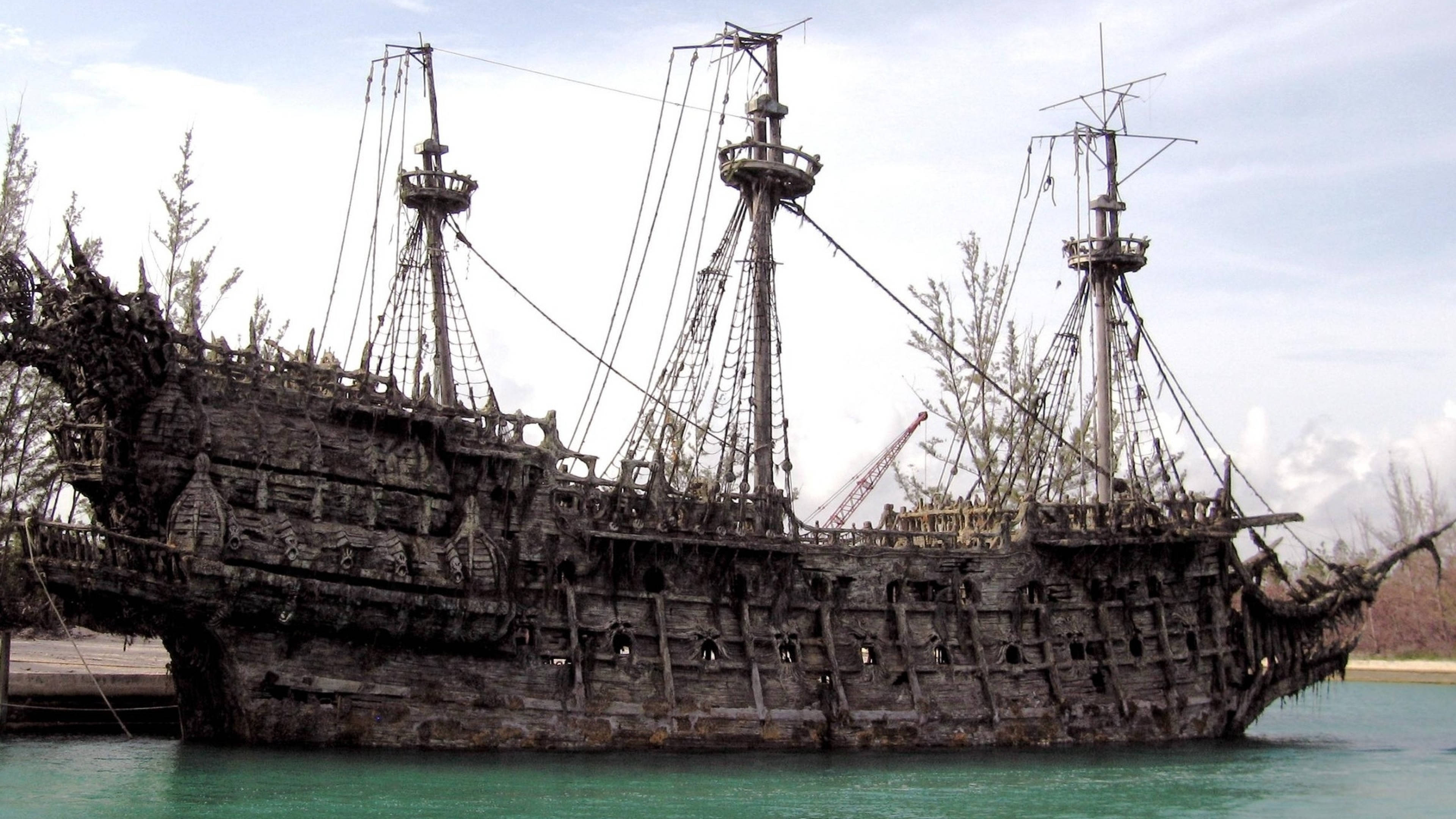 Pirates Of The Caribbean 桌布 壁紙 — 一艘風化的海盜船殘骸擱淺在碧綠色的水中，船體長出樹木，天空多雲。