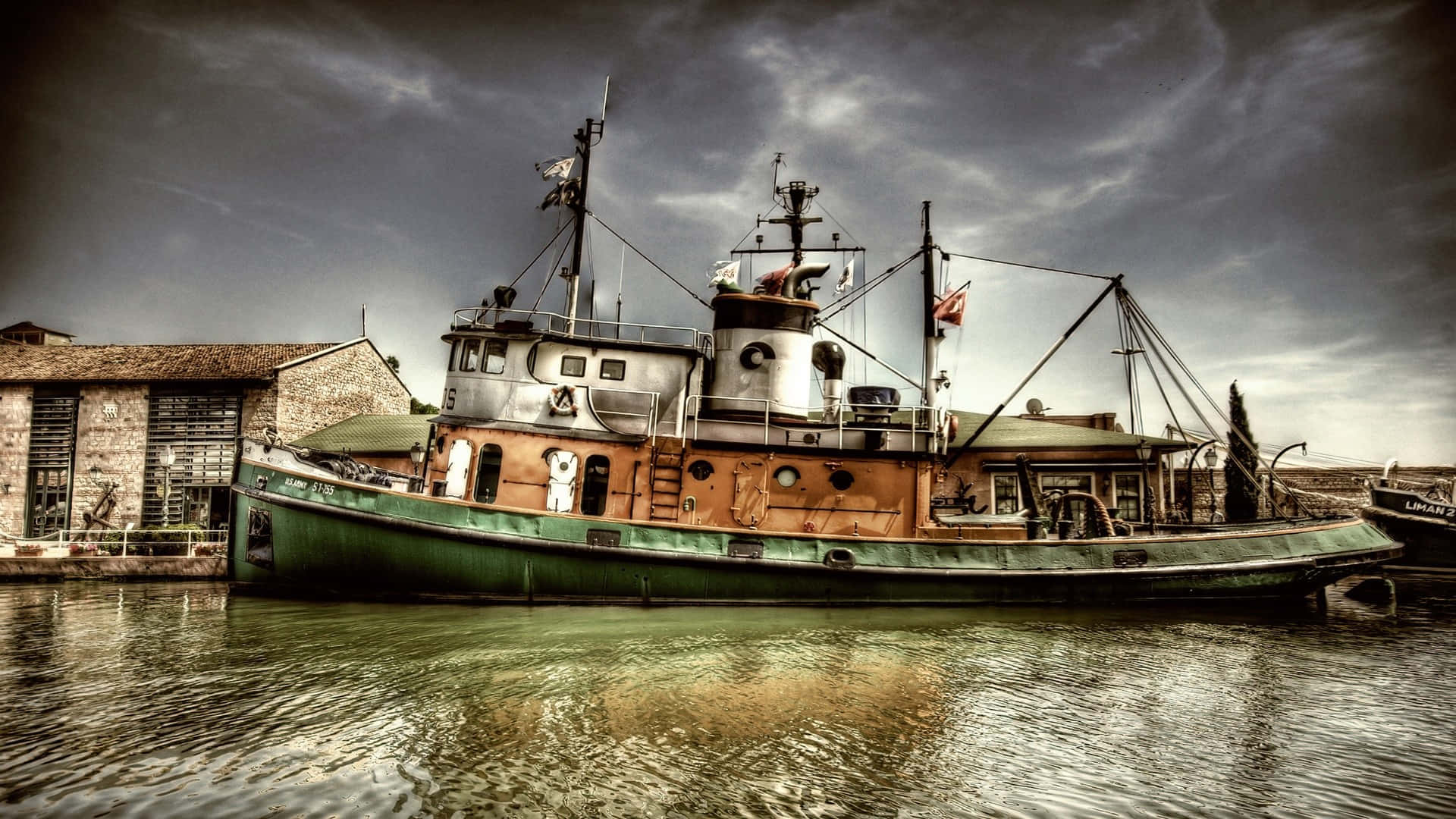 船 桌布 壁紙 — 一艘飽經風霜的綠色和橙色漁船在陰沉多雲的天空下，在石頭建築旁邊的平靜水域中靜止不動。