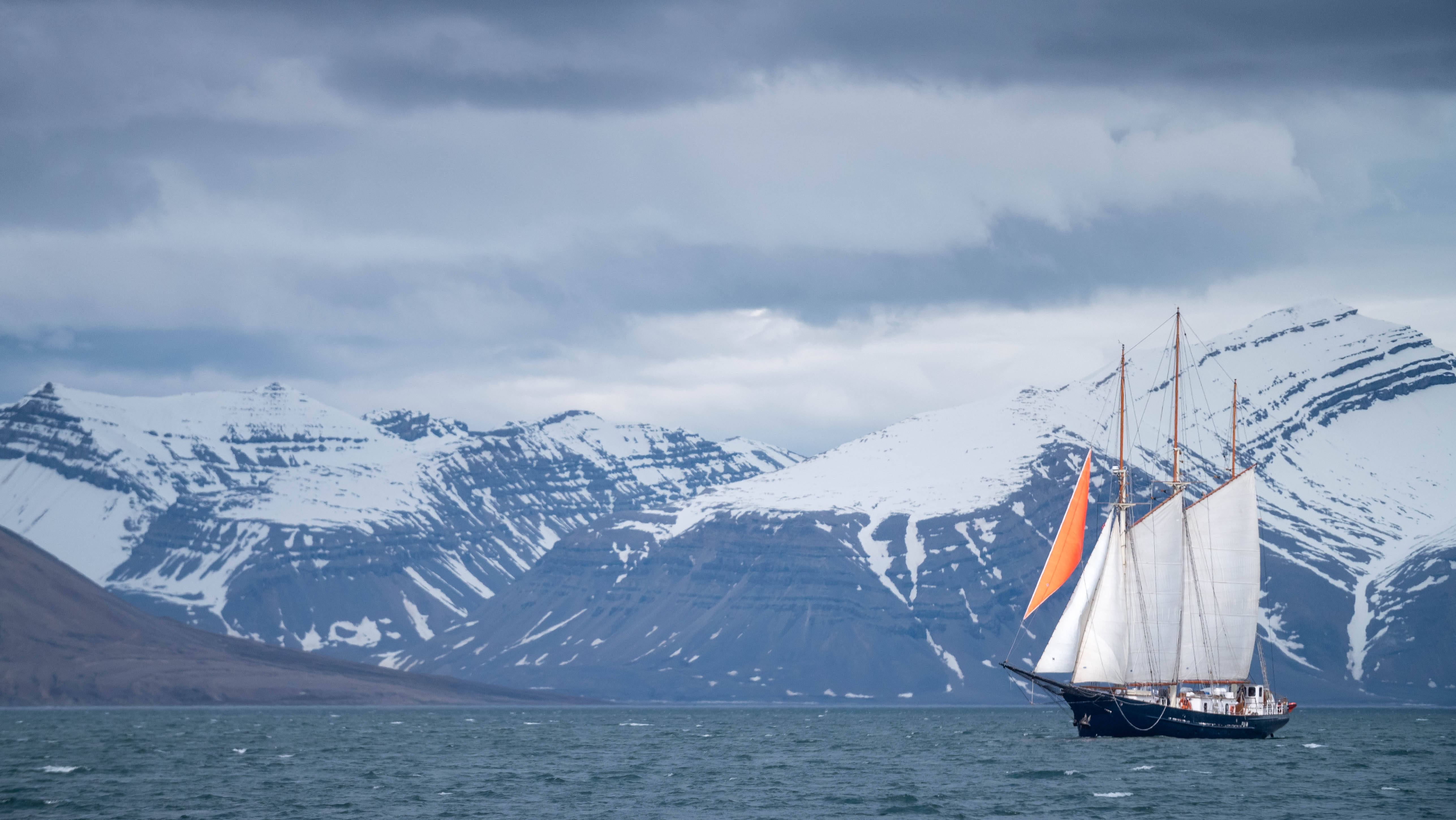 Svalbard 桌布 壁紙 — 一艘高大的帆船，白色帆和一個橙色帆，在波濤洶湧的海面上滑行，前方是白雪覆蓋的山脈，天空多雲。