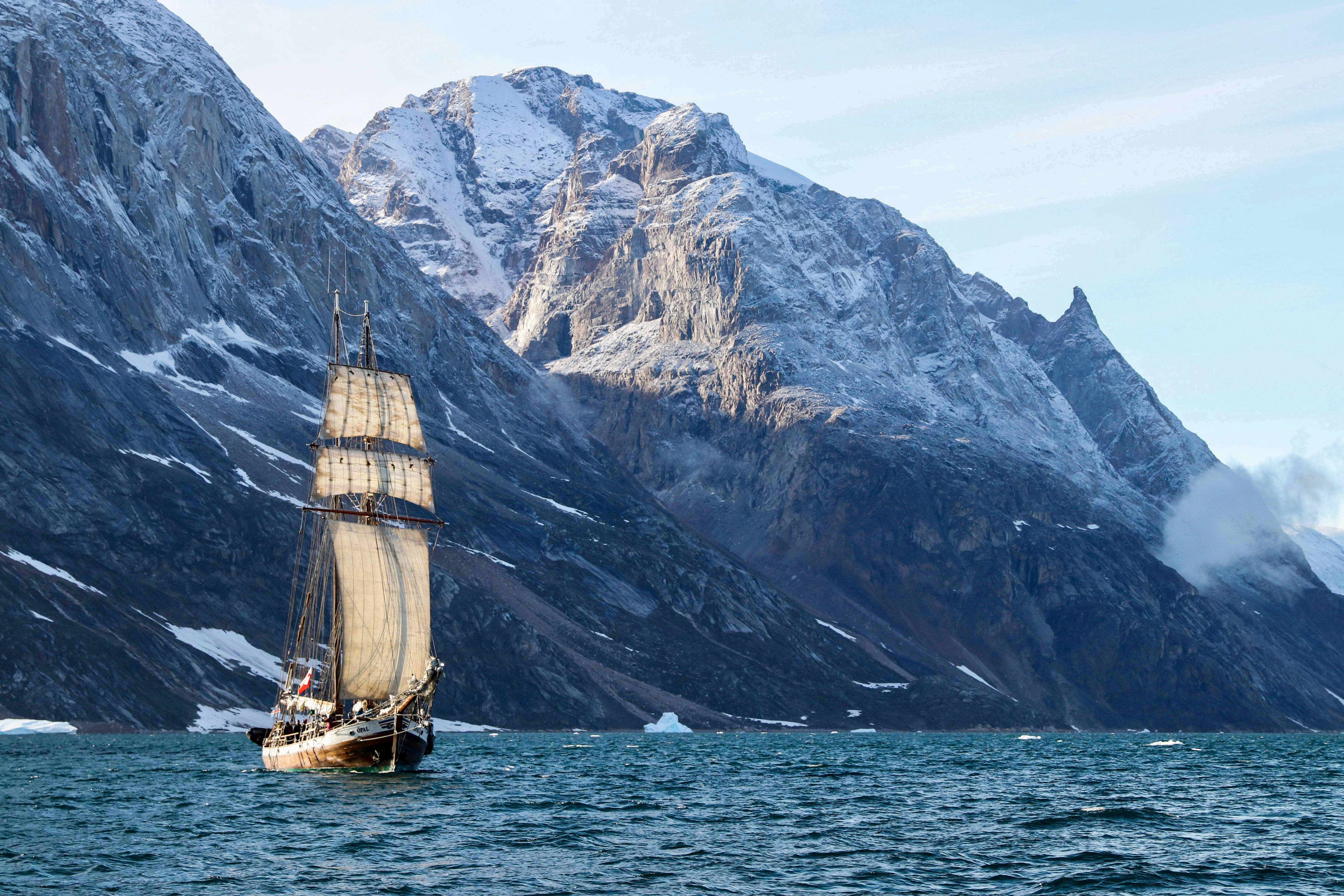 Greenland 桌布 壁紙 — 一艘高大的帆船在冰冷的海水中航行，背景是白雪覆蓋的山脈，氣勢磅礡。