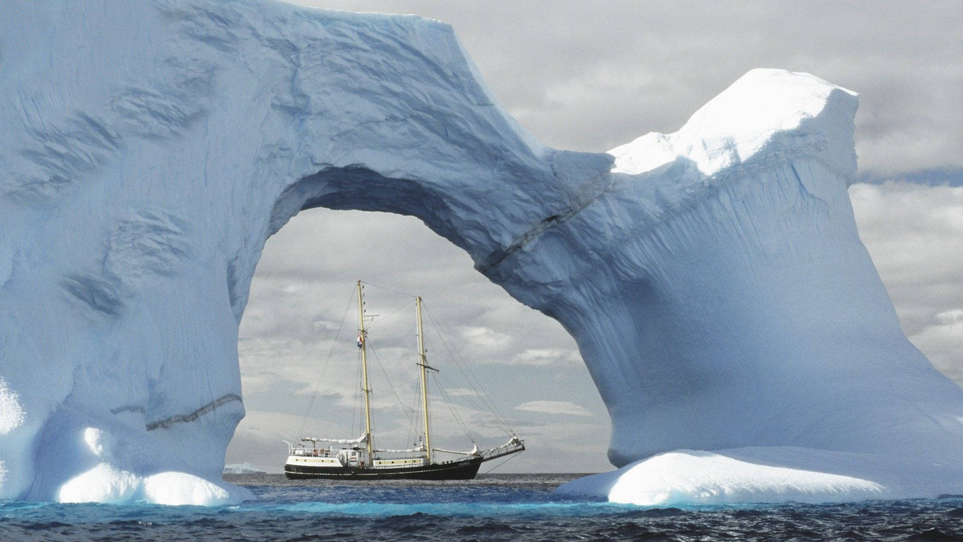Antarctica 桌布 壁紙 — 一艘高大的帆船在多雲的天空下，穿越寒冷的南極海中一個巨大的天然冰拱門。