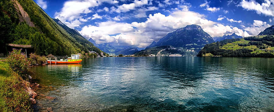 Lake 桌布 壁紙 — 一艘鮮橙色的船隻停泊在寧靜的湖邊，四周是翠綠的山巒，上方是晴朗的藍天和蓬鬆的白雲。