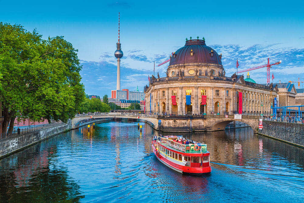 Berlin 桌布 壁紙 — 一艘鮮紅色的觀光船沿著平靜的運河滑行，經過歷史悠久的圓頂建築和茂密的綠樹，在明亮的藍天下，遠處可見柏林電視塔。