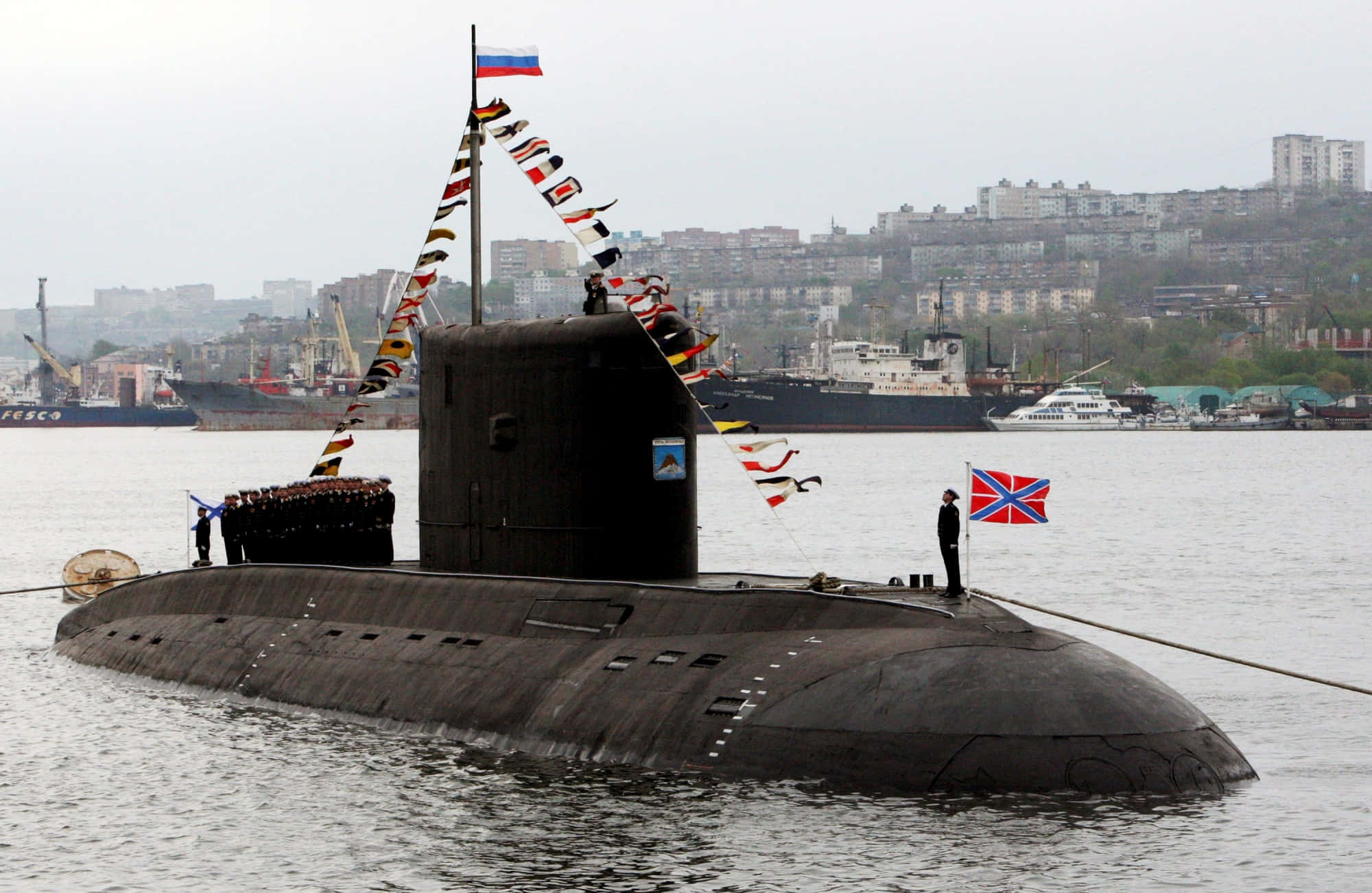 Submarine 桌布 壁紙 — 一艘黑色俄羅斯潛艇在平靜的水面浮出，指揮塔頂有船員和旗幟，背景是城市天際線。