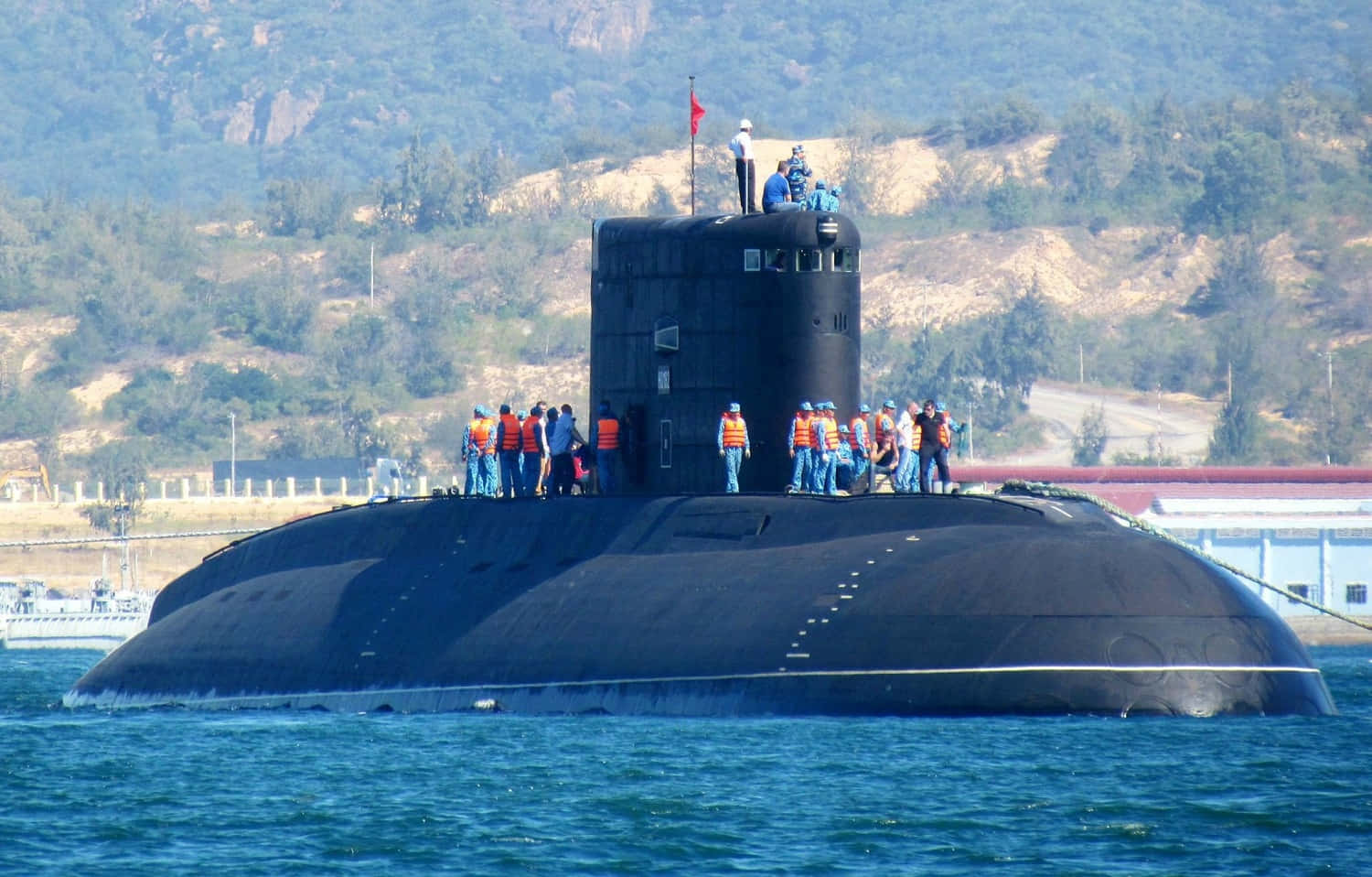 Submarine 桌布 壁紙 — 一艘黑色潛艇在平靜的藍色海水中浮出水面，身穿橙色背心的船員站在甲板上，背景是丘陵海岸線。