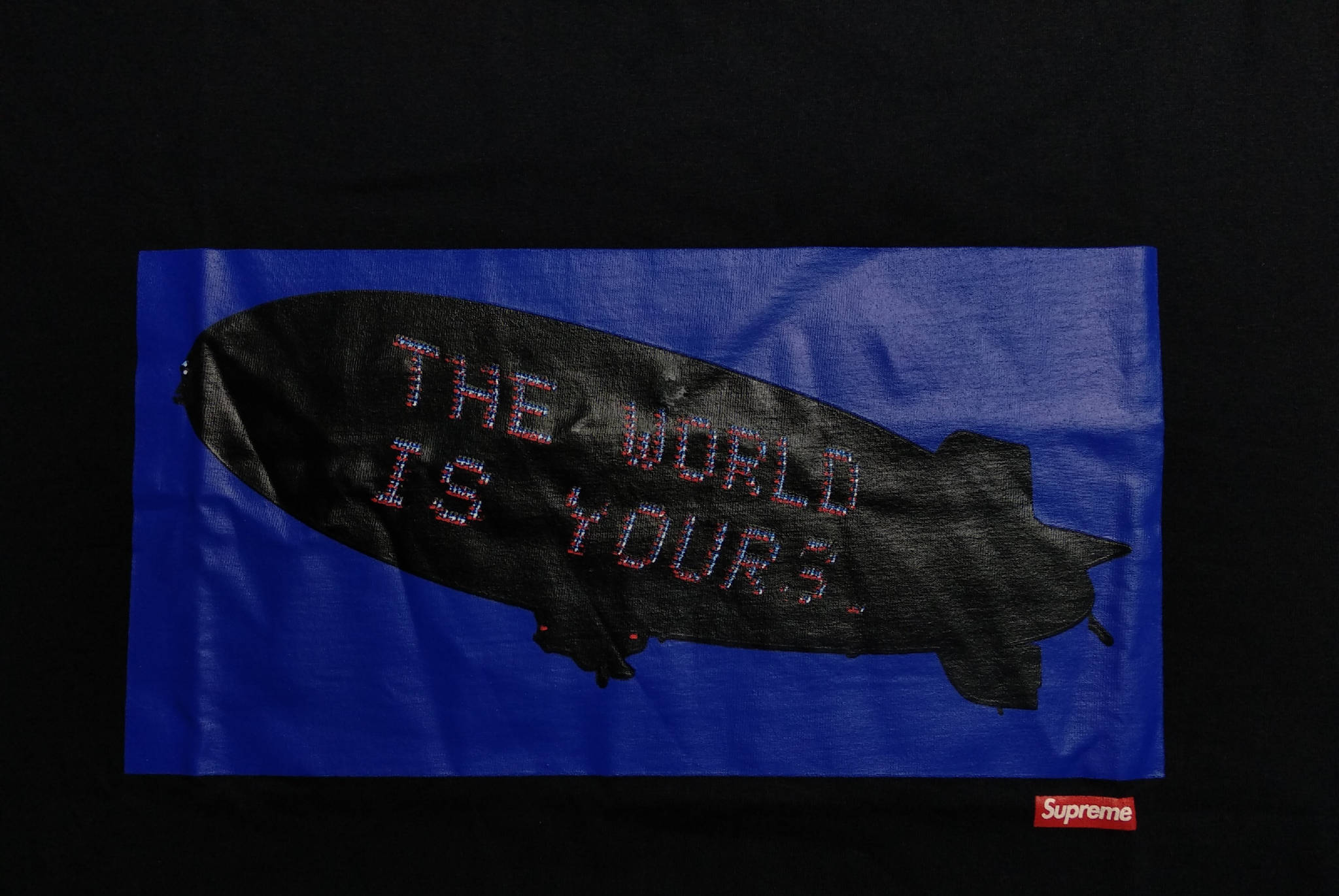 supreme 桌布 壁紙 — 一艘黑色飛船帶有像素化文字 'THE WORLD IS YOURS'，漂浮在鮮豔的藍色背景上，印在帶有紅色 Supreme 標誌的黑色 T 恤上。