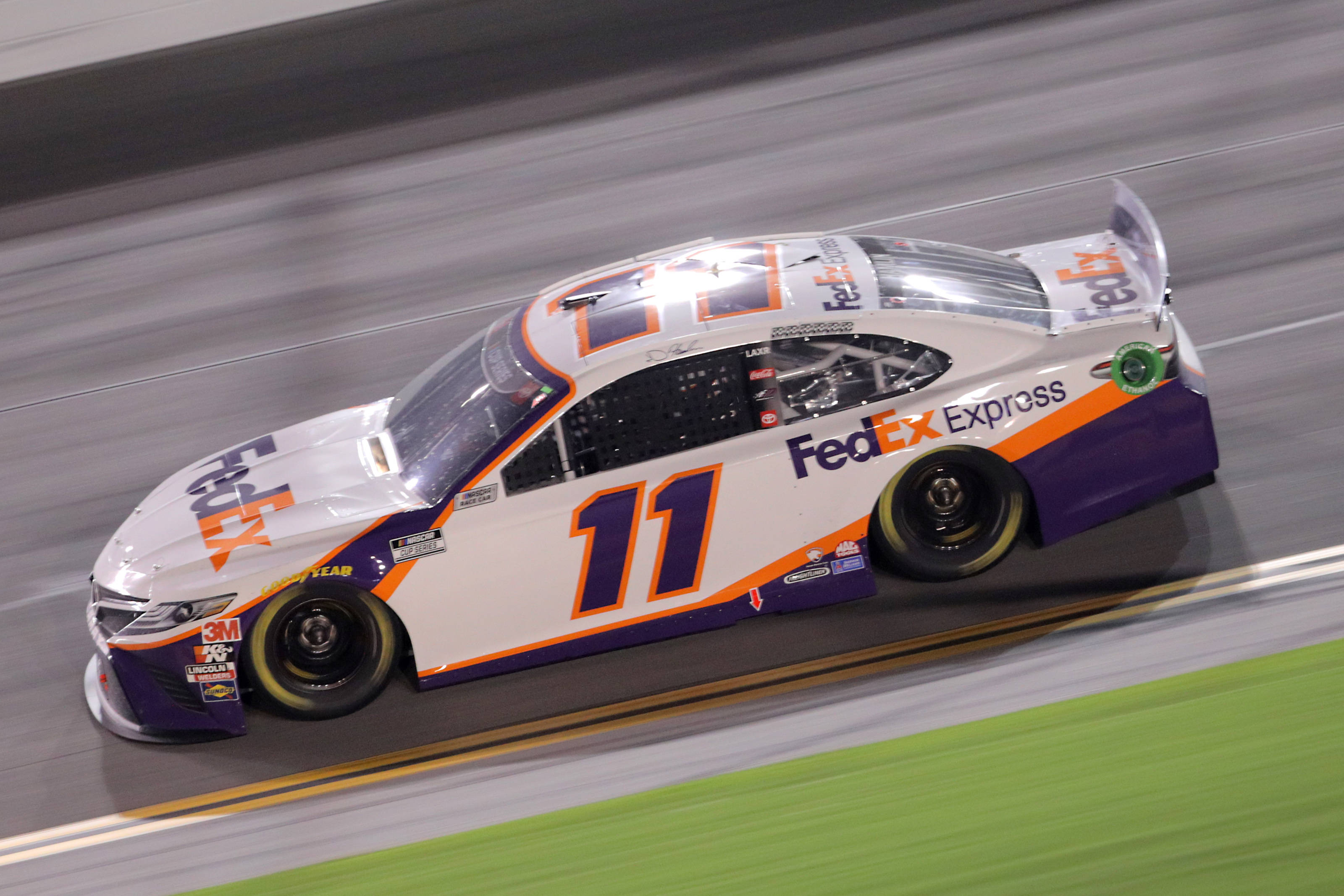 Denny Hamlin 桌布 壁紙 — 一輛 FedEx Express NASCAR 賽車在人造燈光下的賽道上高速駛過，帶有動態模糊效果。