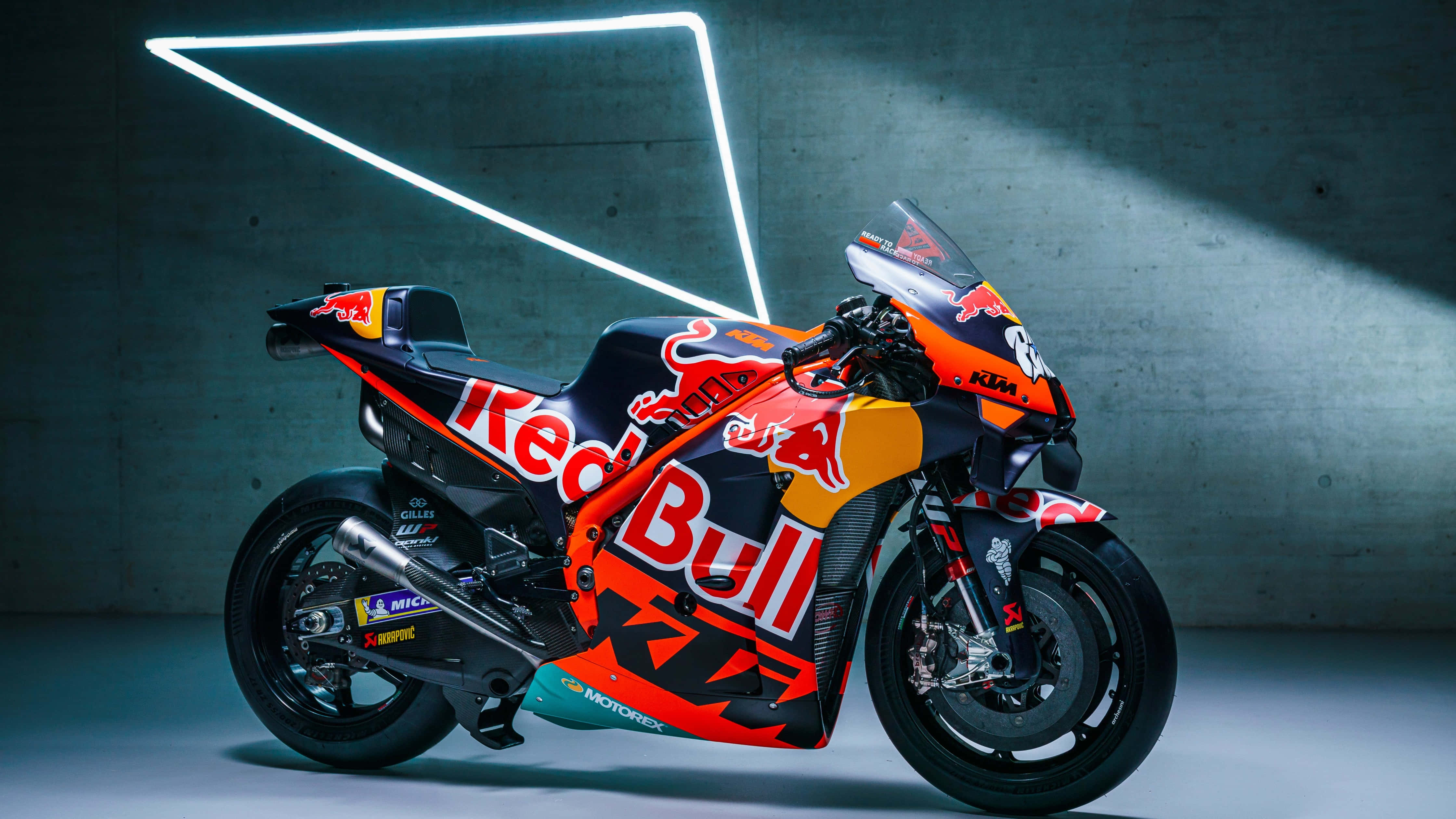 桌布 壁紙 — 一輛 Red Bull KTM 賽車在戲劇性的工作室燈光下，以霓虹三角形裝飾點綴，在混凝土牆前閃閃發光。