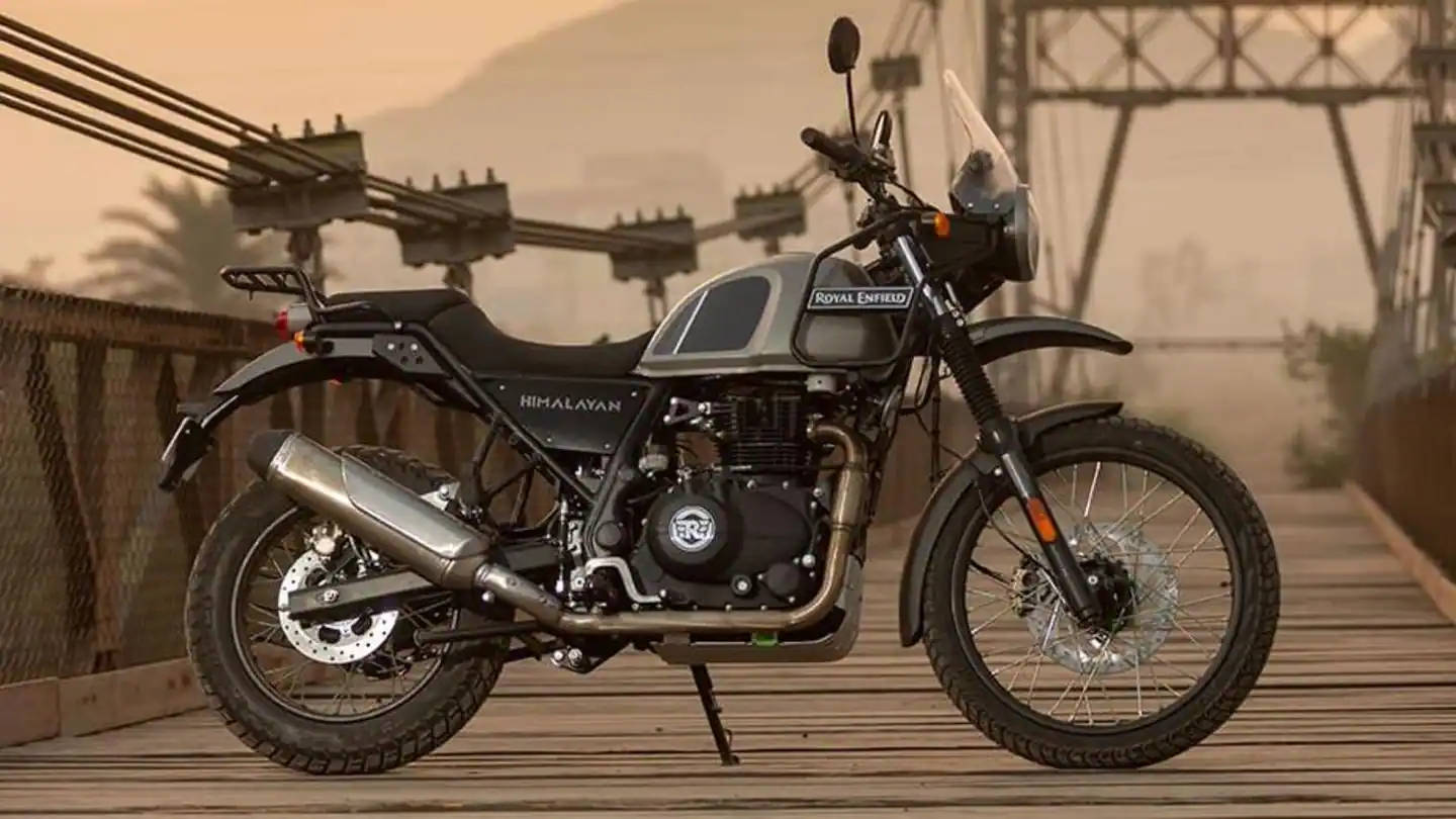  壁紙 — 一輛 Royal Enfield Himalayan 摩托車在朦朧的金色天空下，以遠方的山脈和纜車設施為背景，佇立在木橋上。