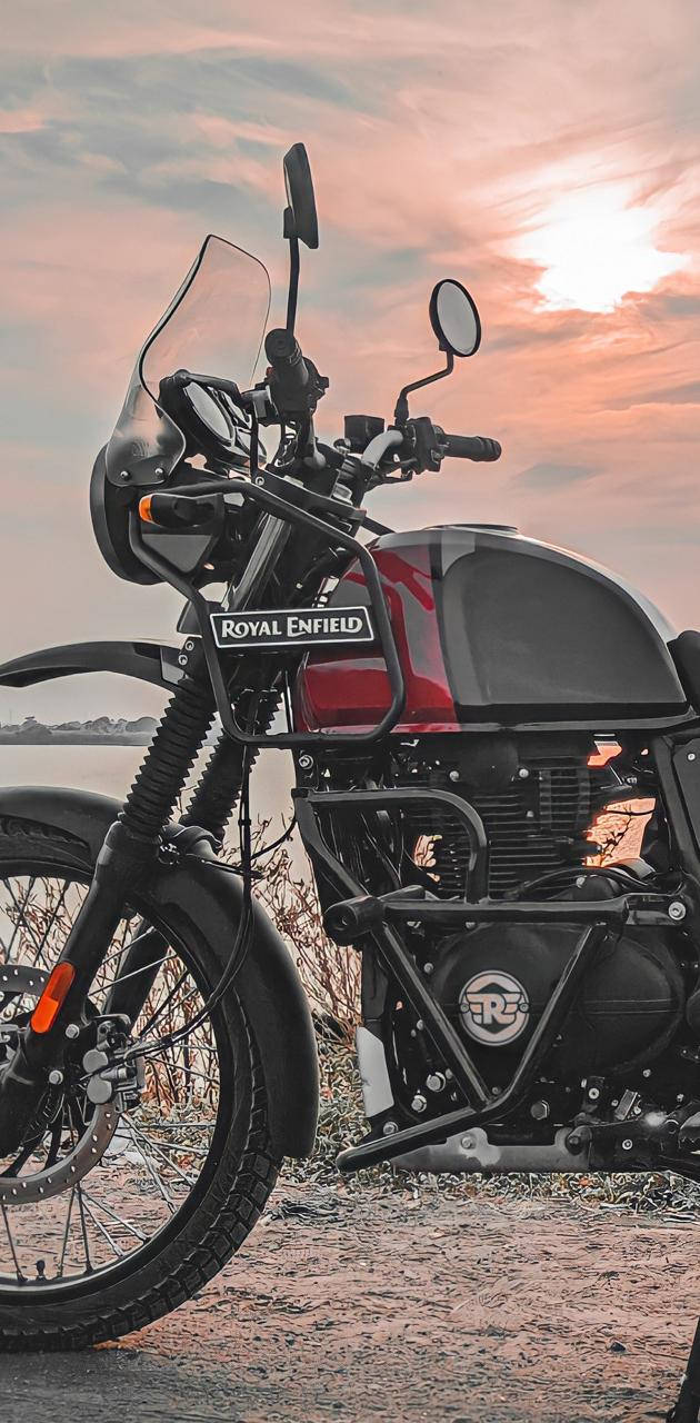腳踏車 桌布 壁紙 — 一輛 Royal Enfield 摩托車在日落時分停在碎石上，背後是溫暖的粉紅色和橙色天空。