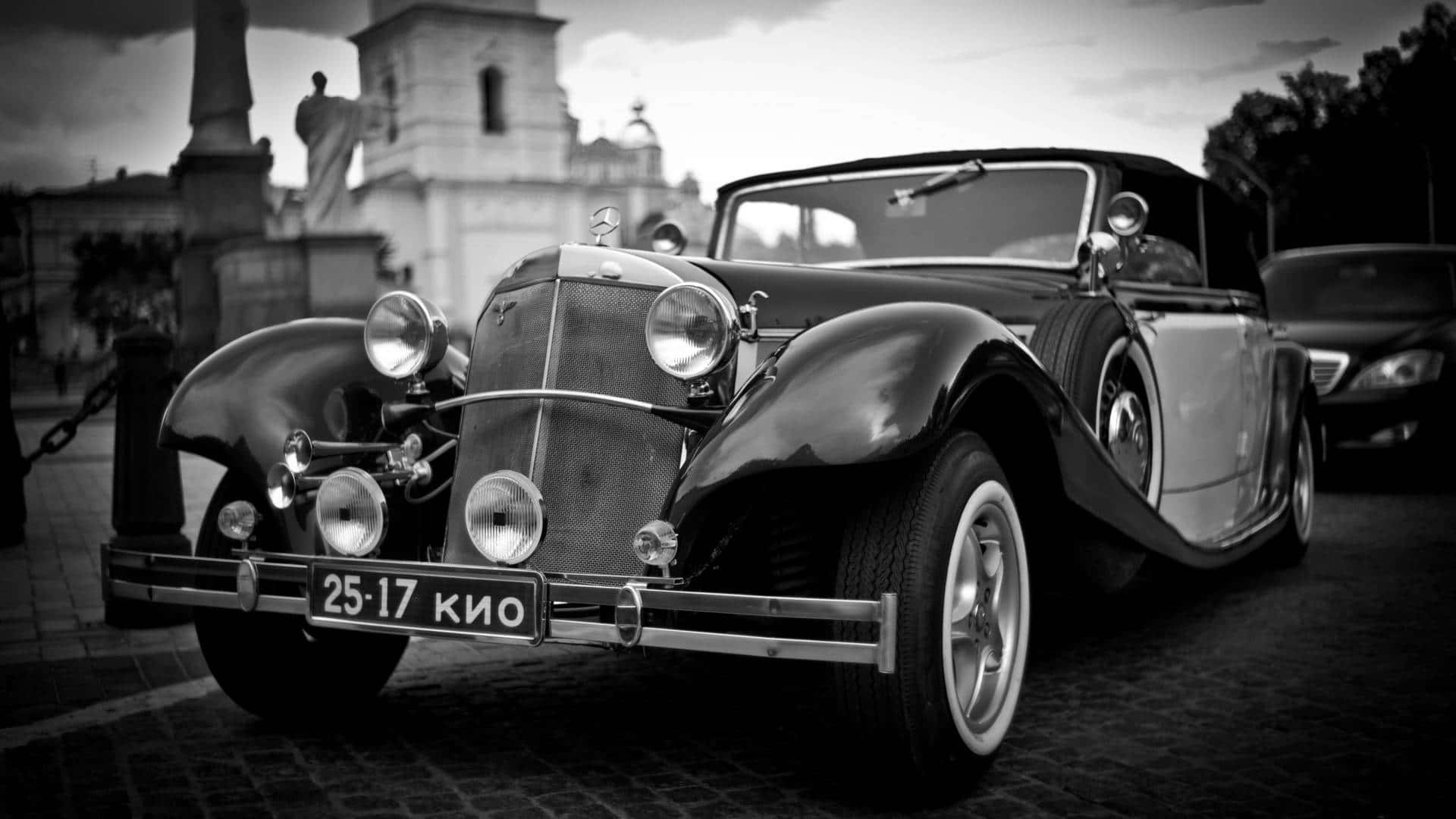 Black And White Vintage Car 桌布 壁紙 — 一輛 vintage Mercedes-Benz 敞篷車停泊在鵝卵石街道上，背景是歷史建築。