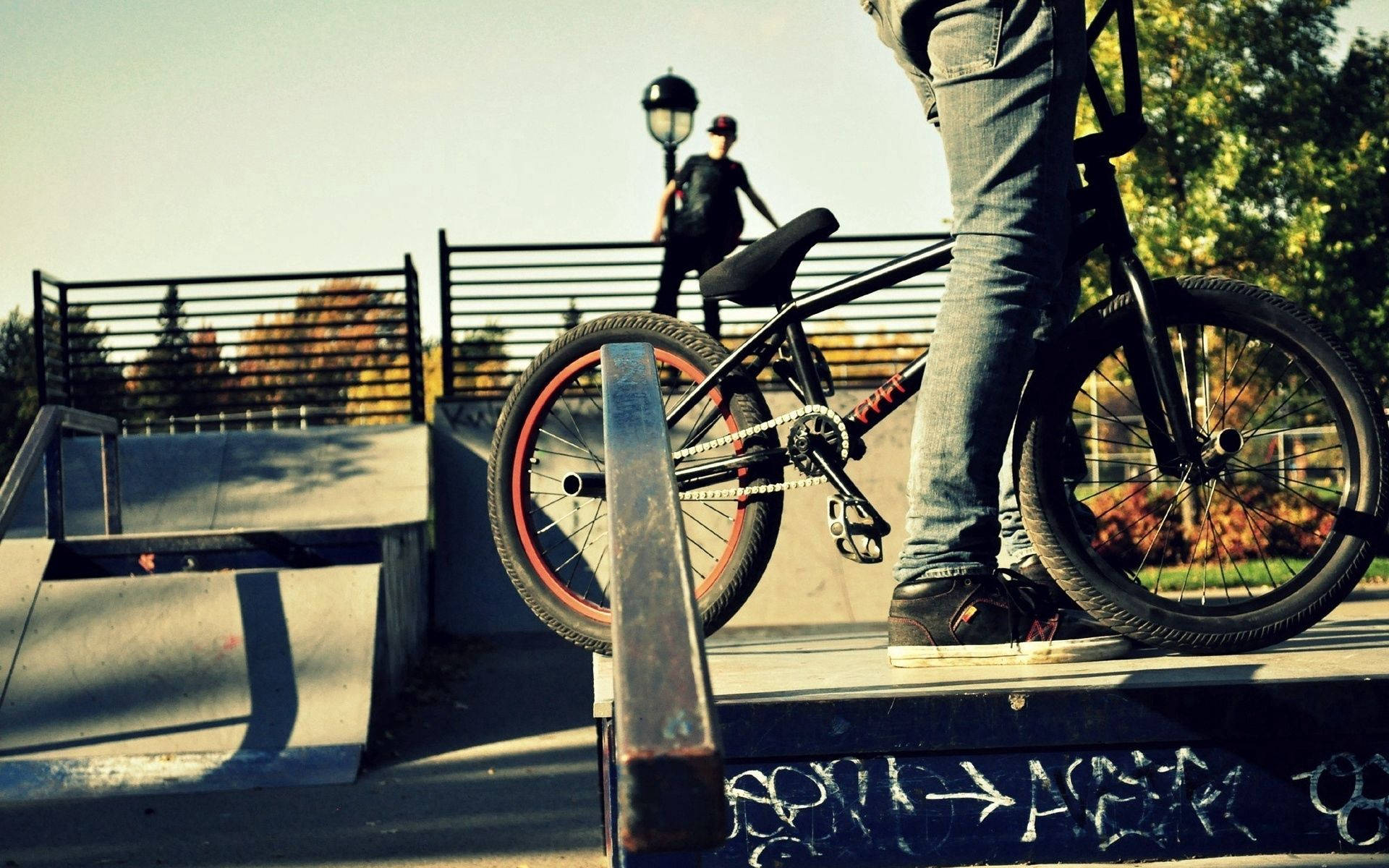 人物 桌布 壁紙 — 一輛BMX腳踏車靠在滑板公園的欄杆上，一名車手站在附近，沐浴在溫暖的秋日陽光下。