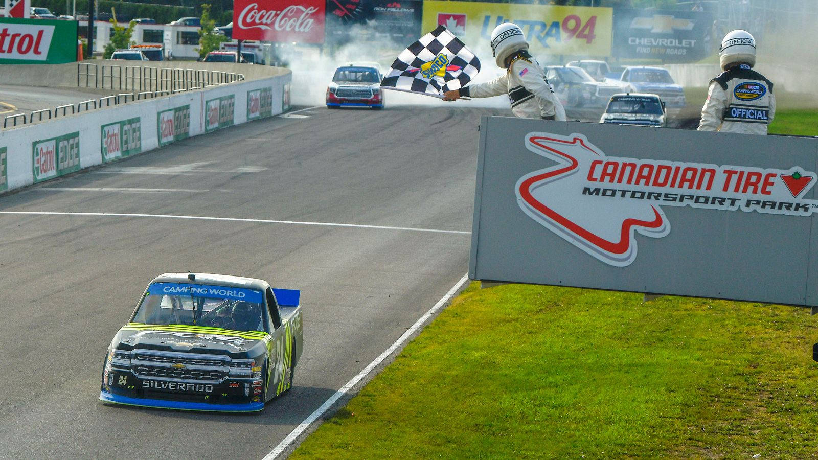 Racing 桌布 壁紙 — 一輛NASCAR卡車在加拿大輪胎賽車公園衝過終點線，工作人員在賽車煙霧中揮舞著方格旗。