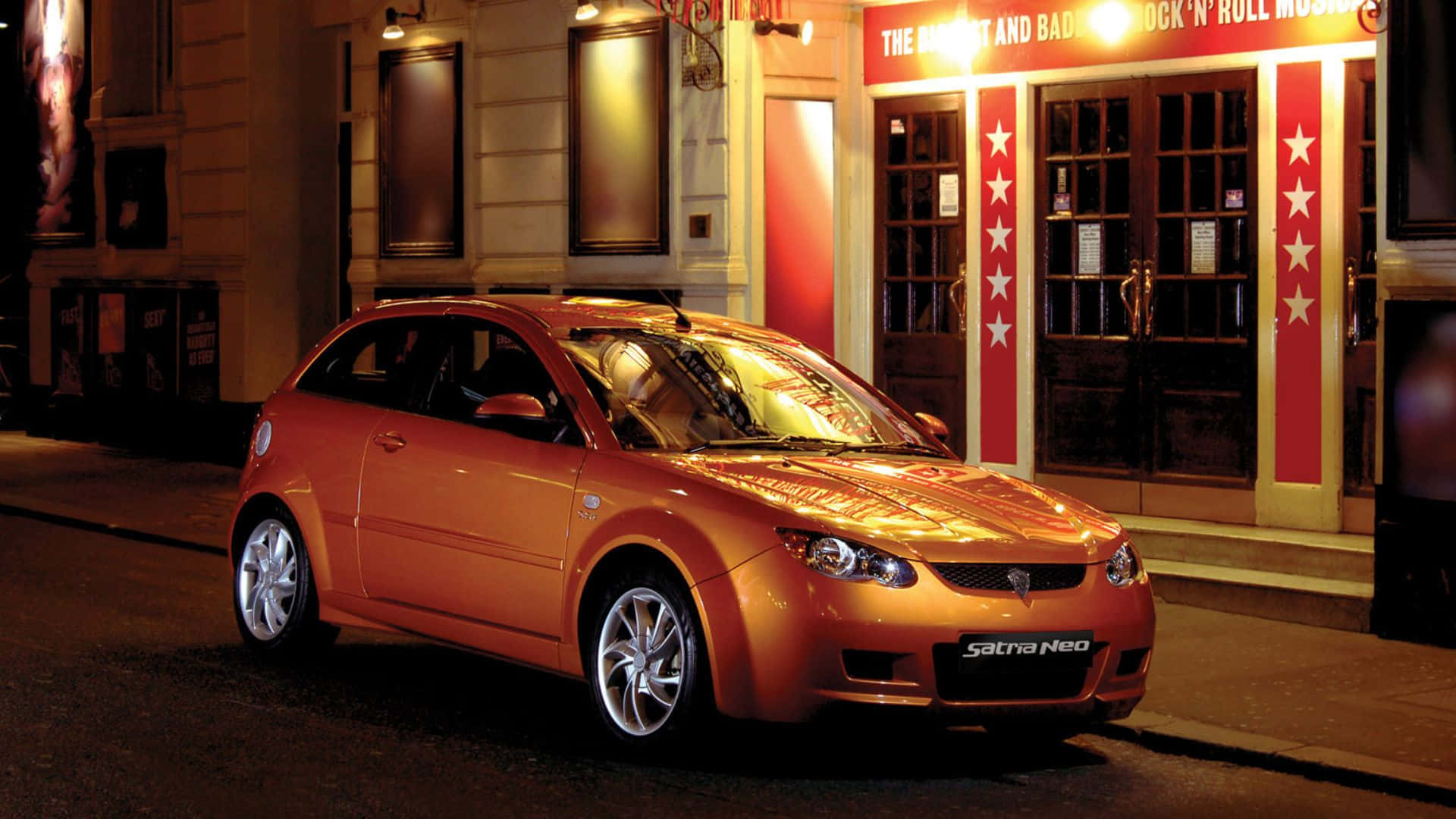 Proton 桌布 壁紙 — 一輛亮橙色 Saturn Neo 掀背車在夜間停放在一個燈火通明、帶有紅色星柱的搖滾博物館前。