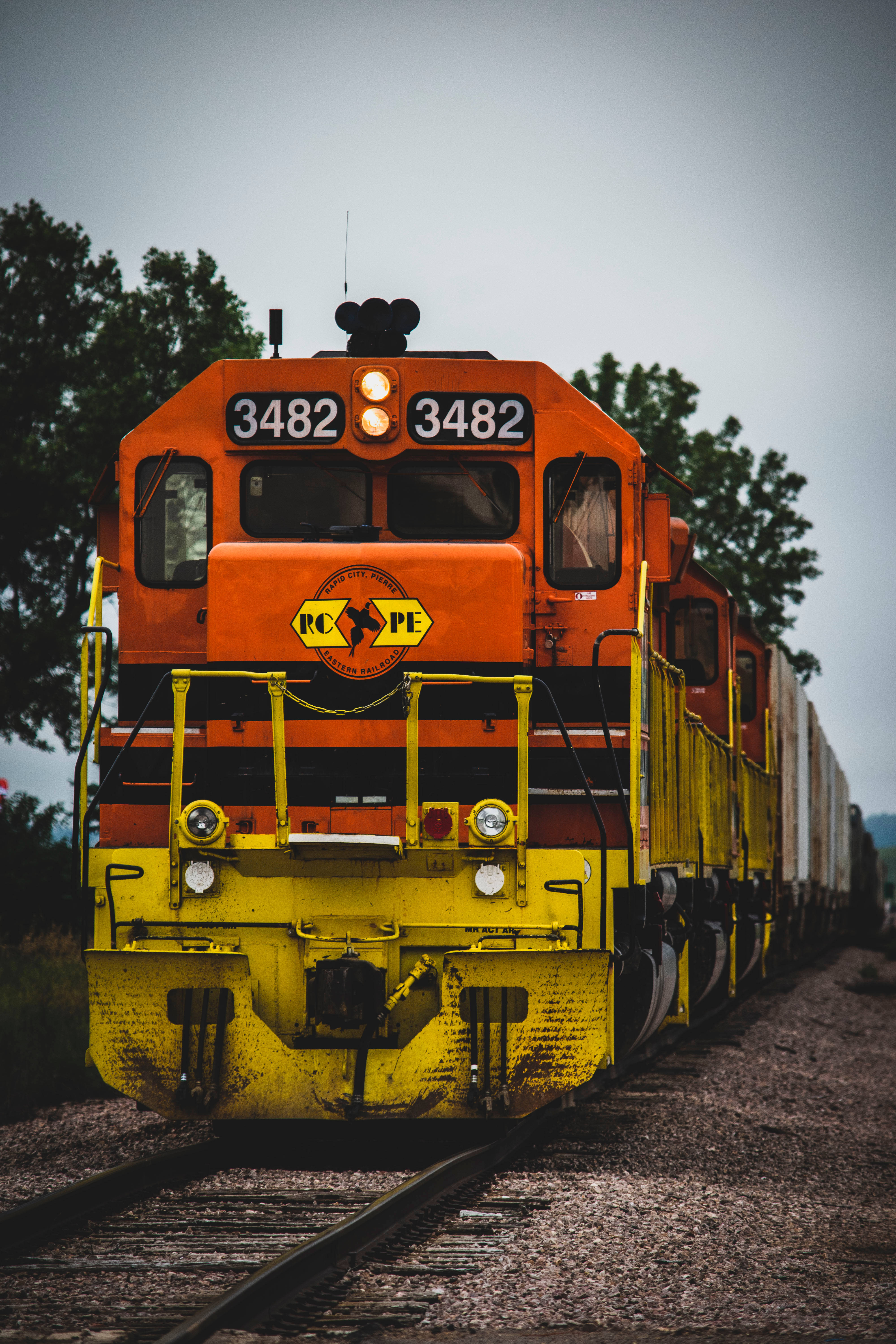 火車 桌布 壁紙 — 一輛亮橙色和黃色的貨運火車引擎編號3482，在陰天背景下，停在有樹木的軌道上。