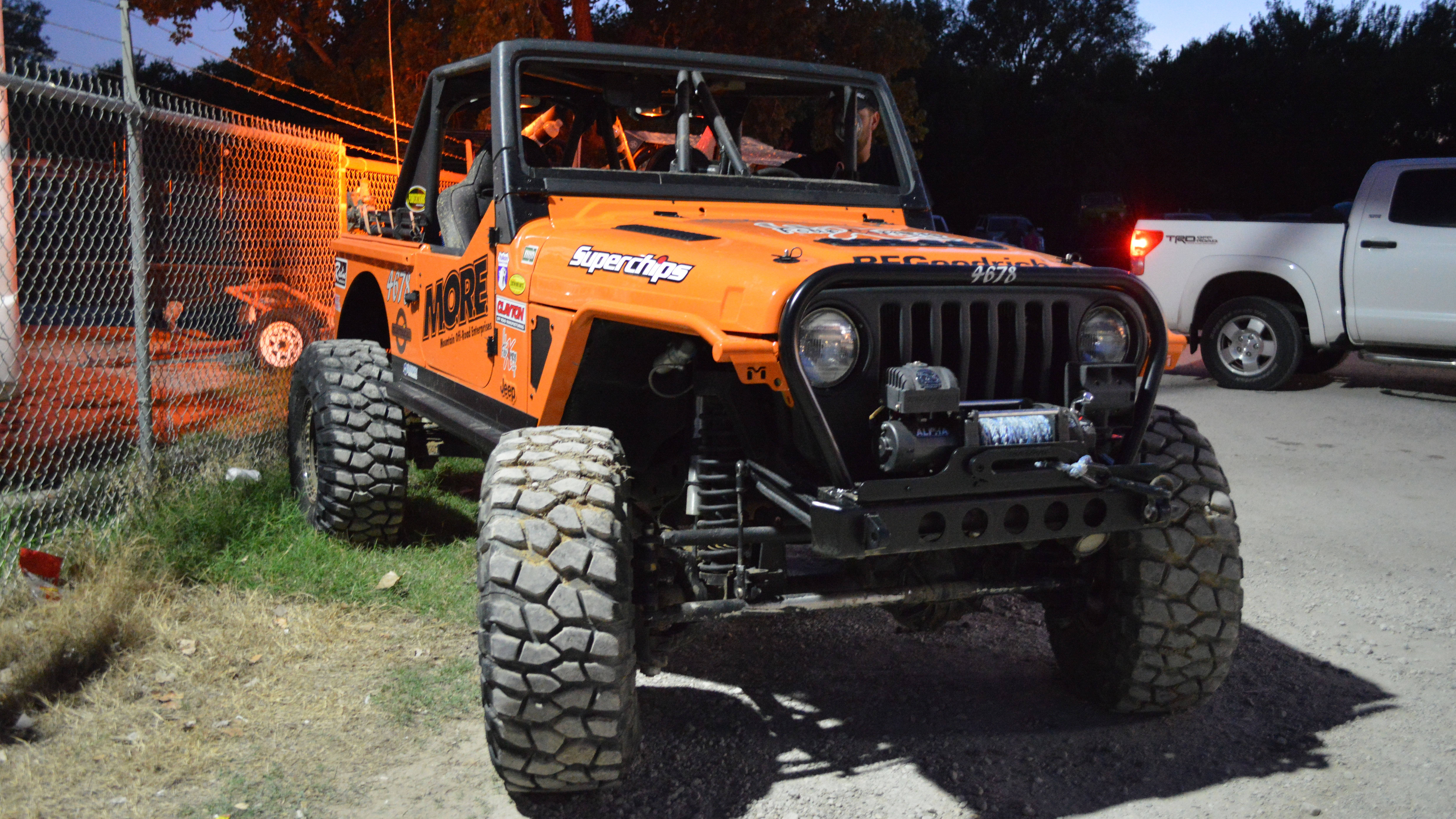 汽車 桌布 壁紙 — 一輛亮橙色改裝Jeep Wrangler，配備超大輪胎和賽車貼紙，在黃昏時停泊在鏈條圍欄附近的碎石地上。