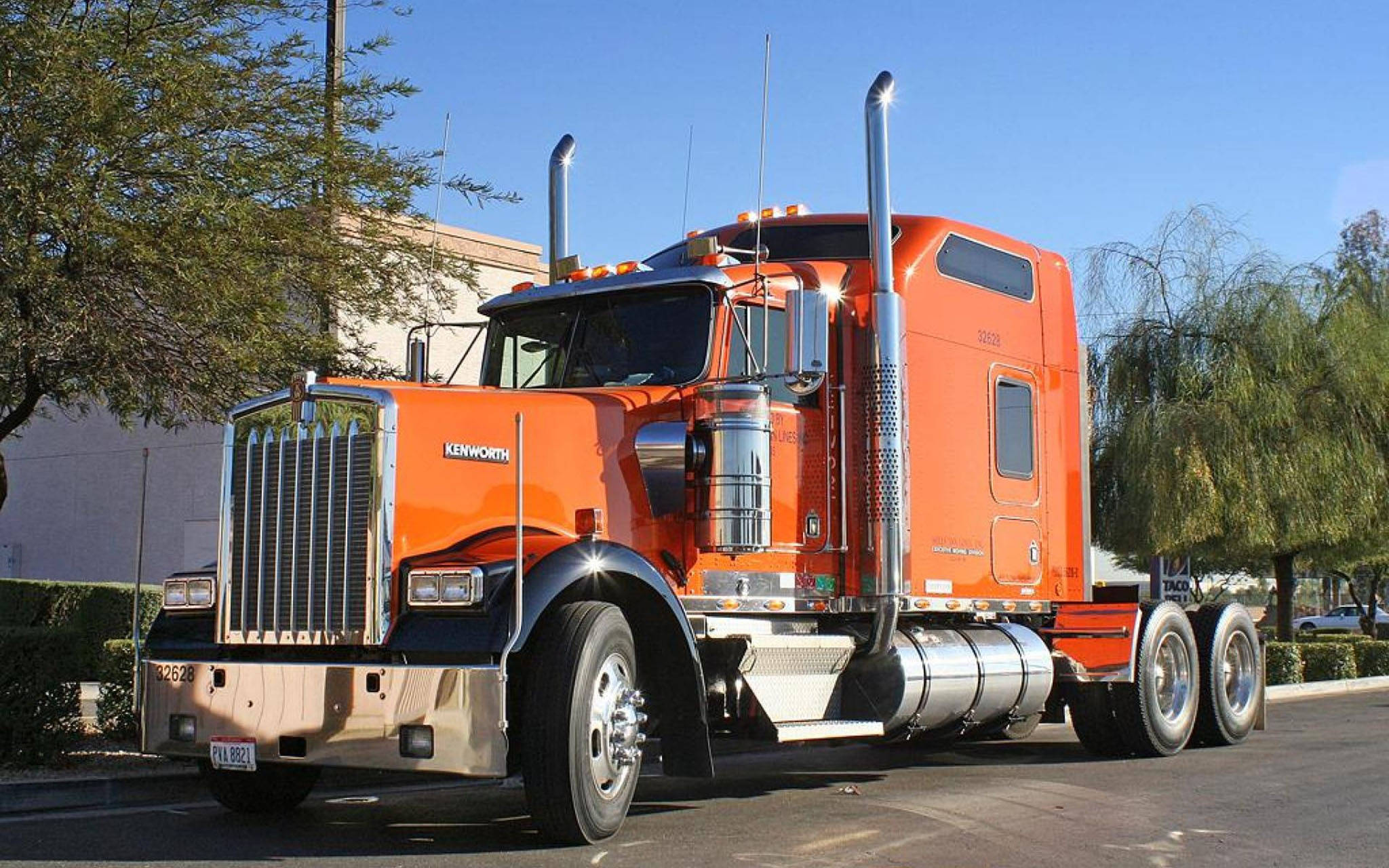 Kenworth W900 桌布 壁紙 — 一輛亮橙色的Kenworth半掛卡車在晴朗的藍天下，停泊在陽光普照、綠樹成蔭的街道上閃閃發光。
