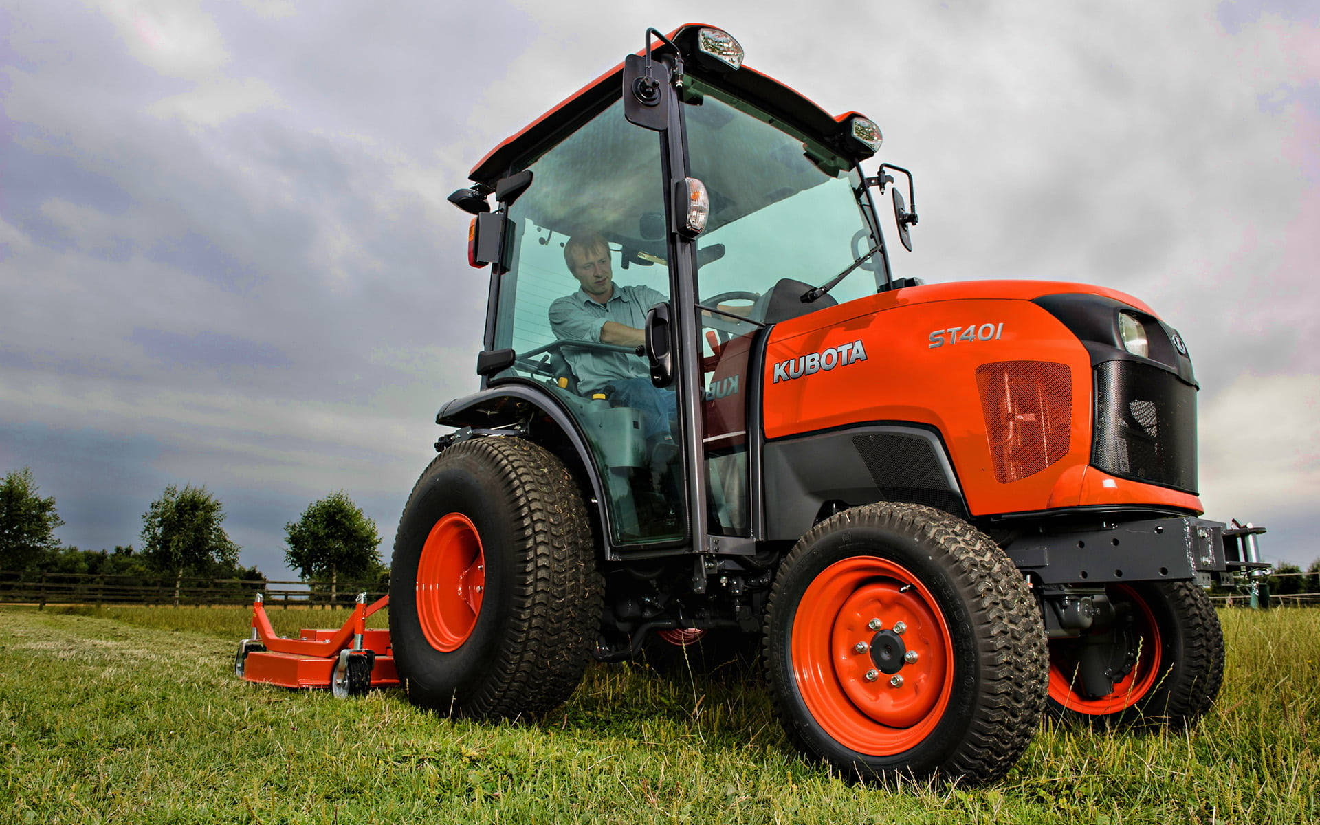 Farmer 桌布 壁紙 — 一輛亮橙色的Kubota ST401拖拉機，內有農夫，停泊在陰天下的草地上。