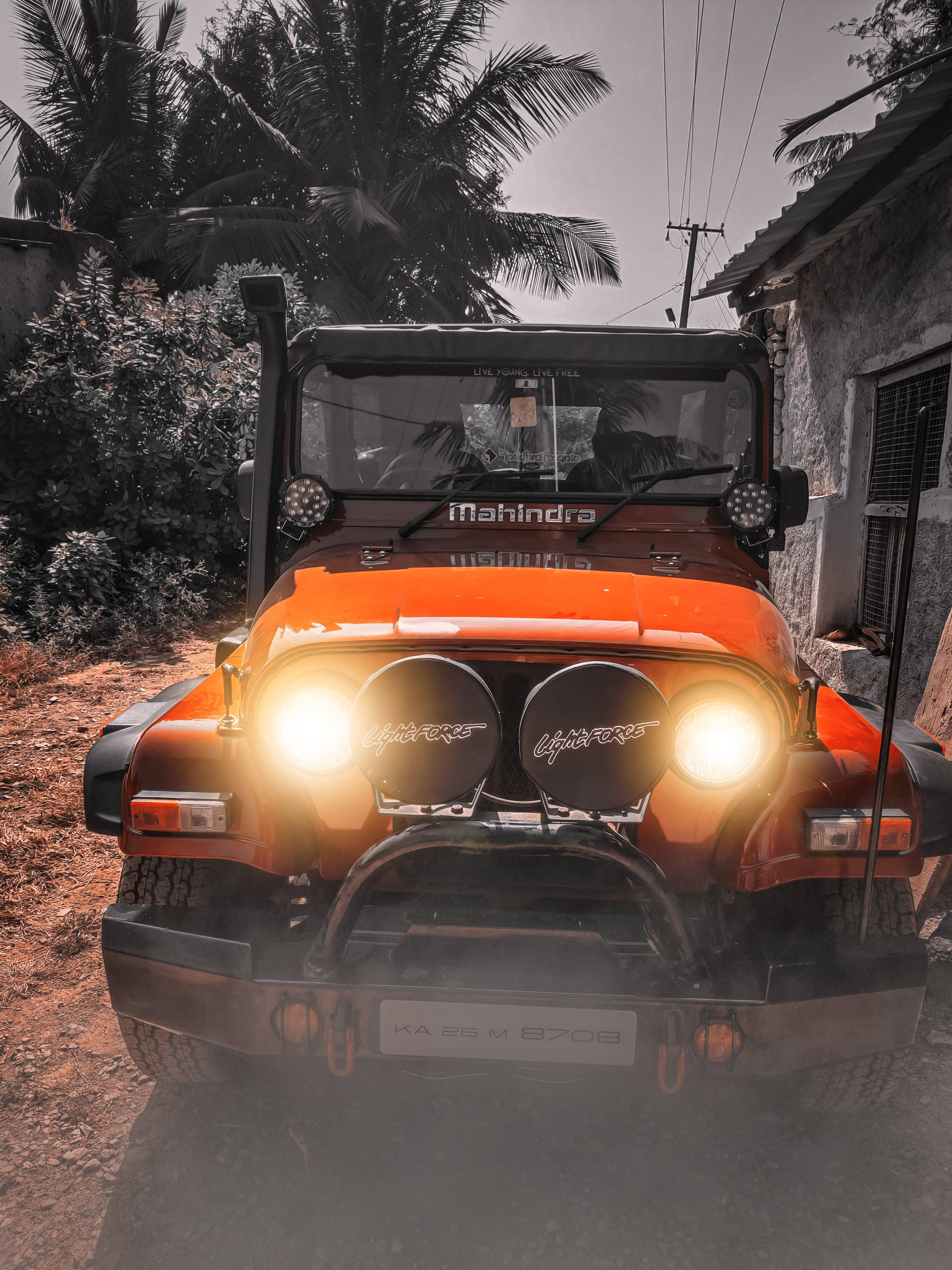 人像動態 桌布 壁紙 — 一輛亮橙色的Mahindra Thar吉普車，車頭燈閃爍，停泊在棕櫚樹下的塵土鄉村道路上。