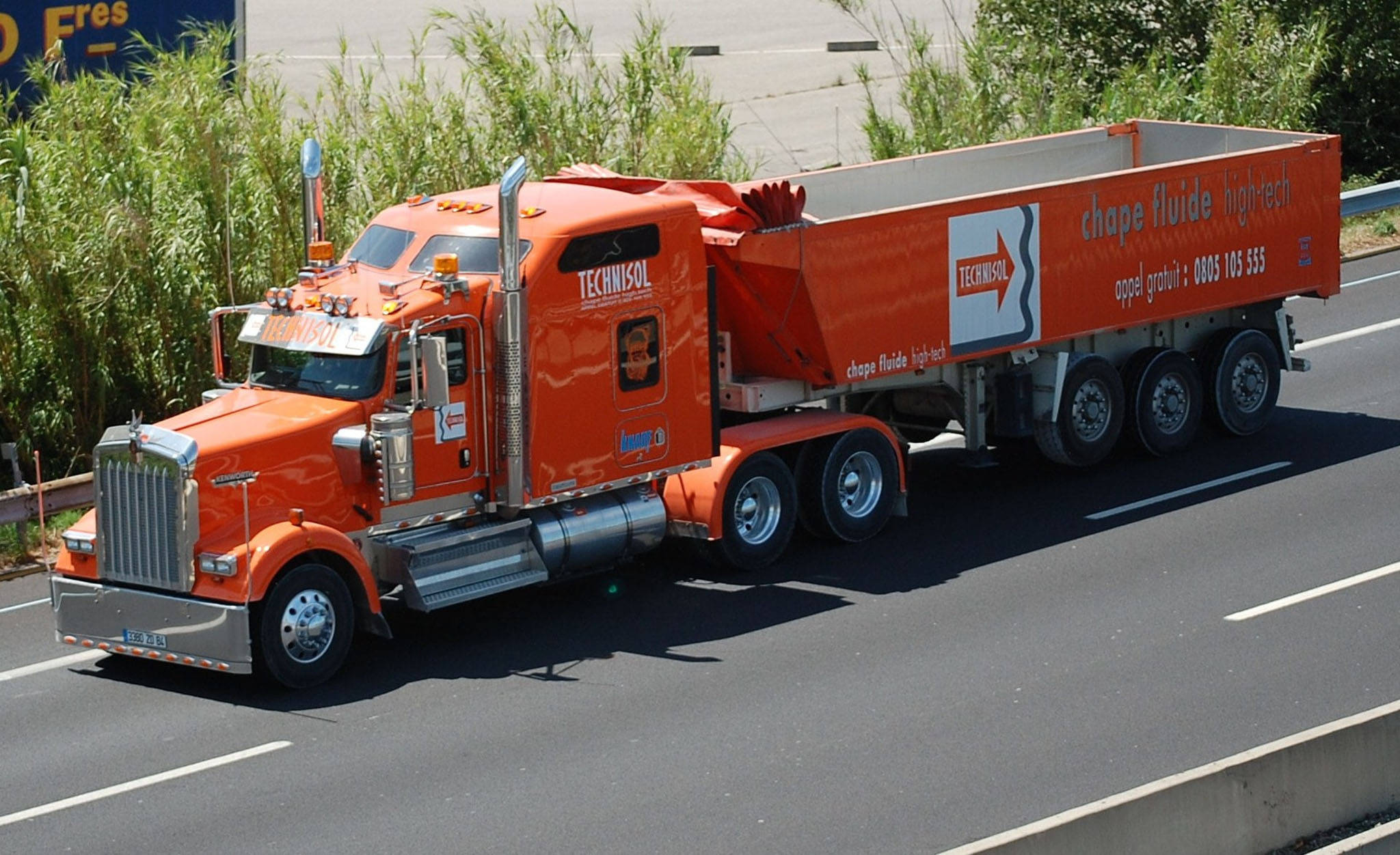 Kenworth W900 桌布 壁紙 — 一輛亮橙色的Technisol混凝土攪拌車在陽光明媚的高速公路上行駛，兩旁是高高的綠草。