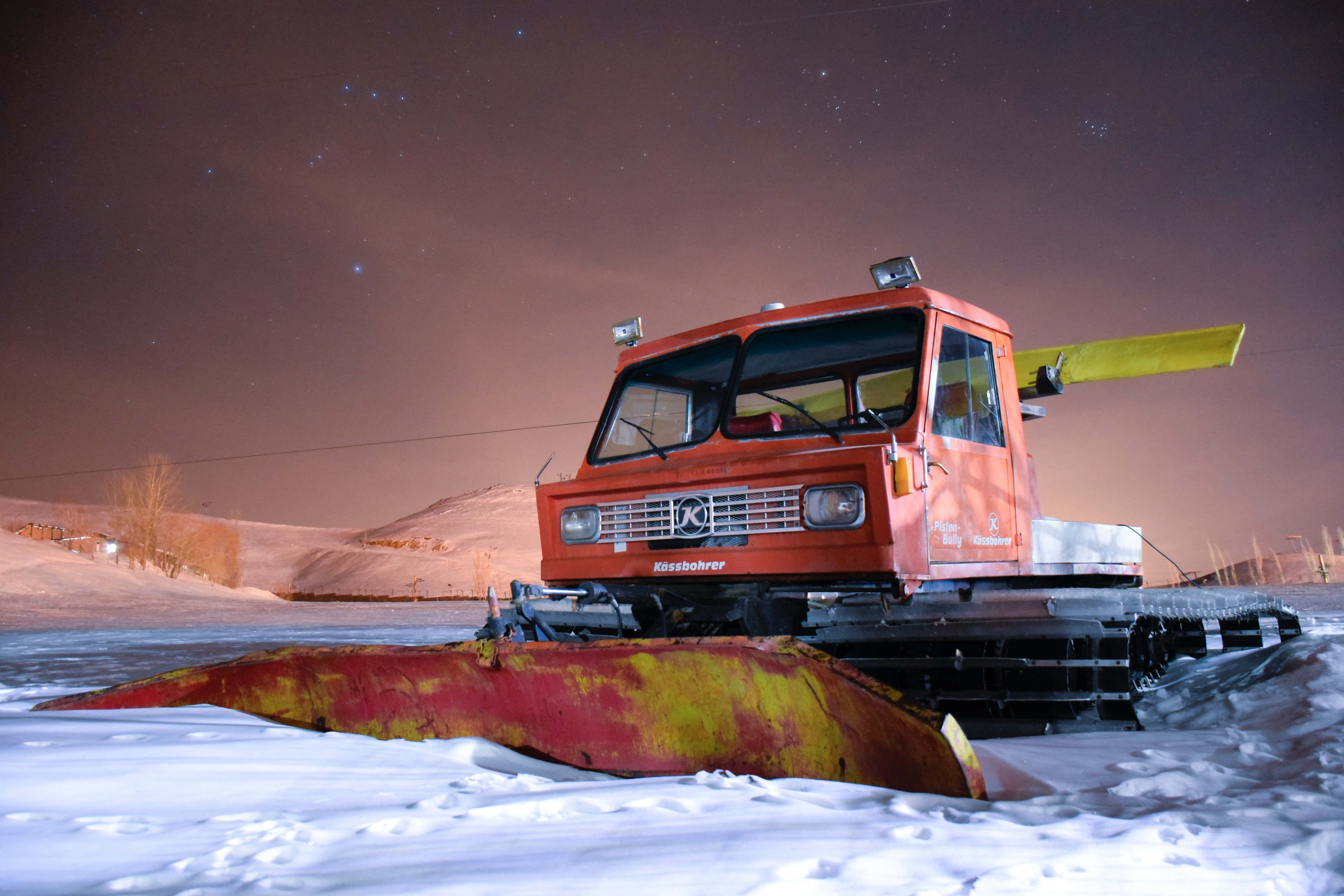 Snowplow 桌布 壁紙 — 一輛亮橙色的掃雪車停在結冰的湖面上，背景是星空和白雪覆蓋的山丘。
