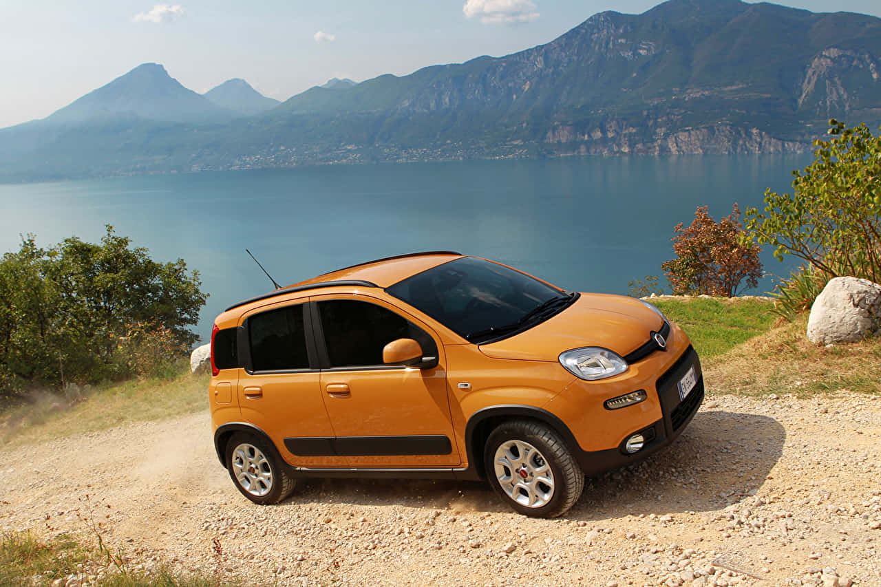 Fiat Panda 桌布 壁紙 — 一輛亮橙色的緊湊型汽車停在碎石觀景點上，背景是廣闊的藍色湖泊和山脈，天空晴朗。